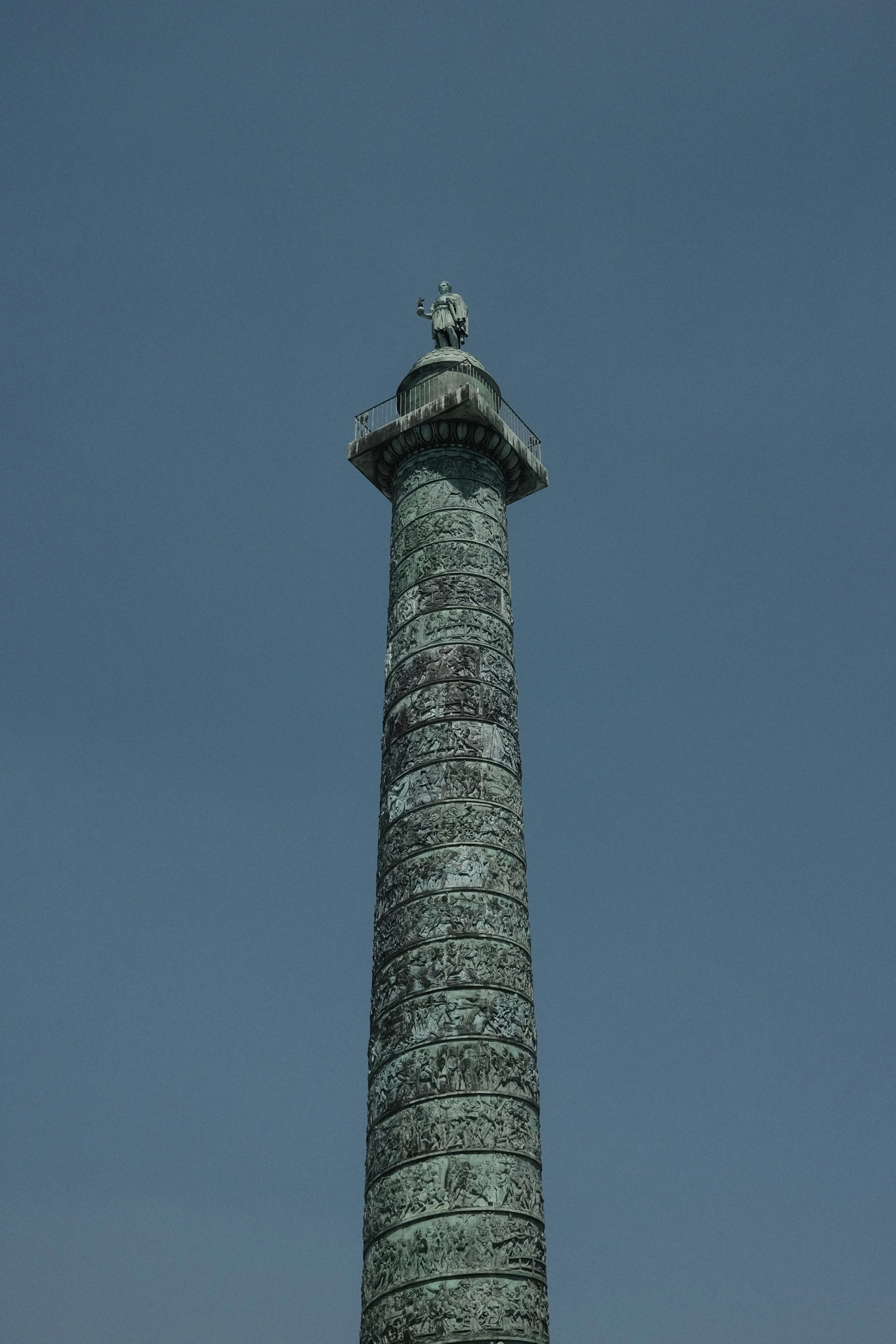 A tall cylindrical monument with intricate carvings and a statue of a man at the top, standing against a clear blue sky.