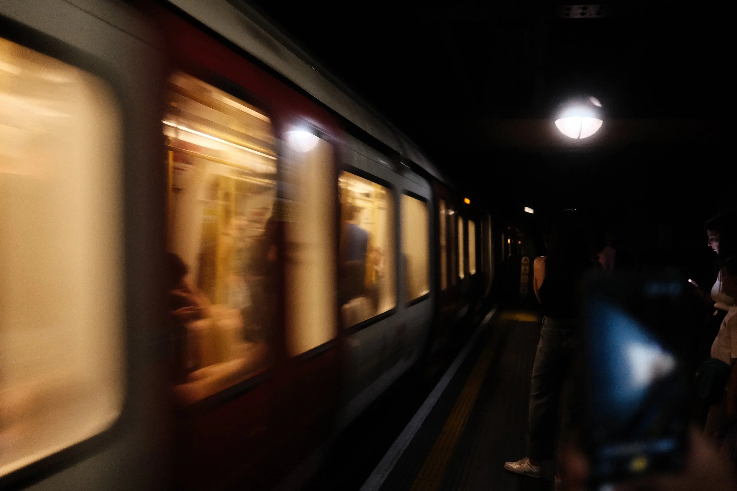 A night scene of a subway train with illuminated windows at a station platform, with some people standing nearby, and a ceiling light visible above.
