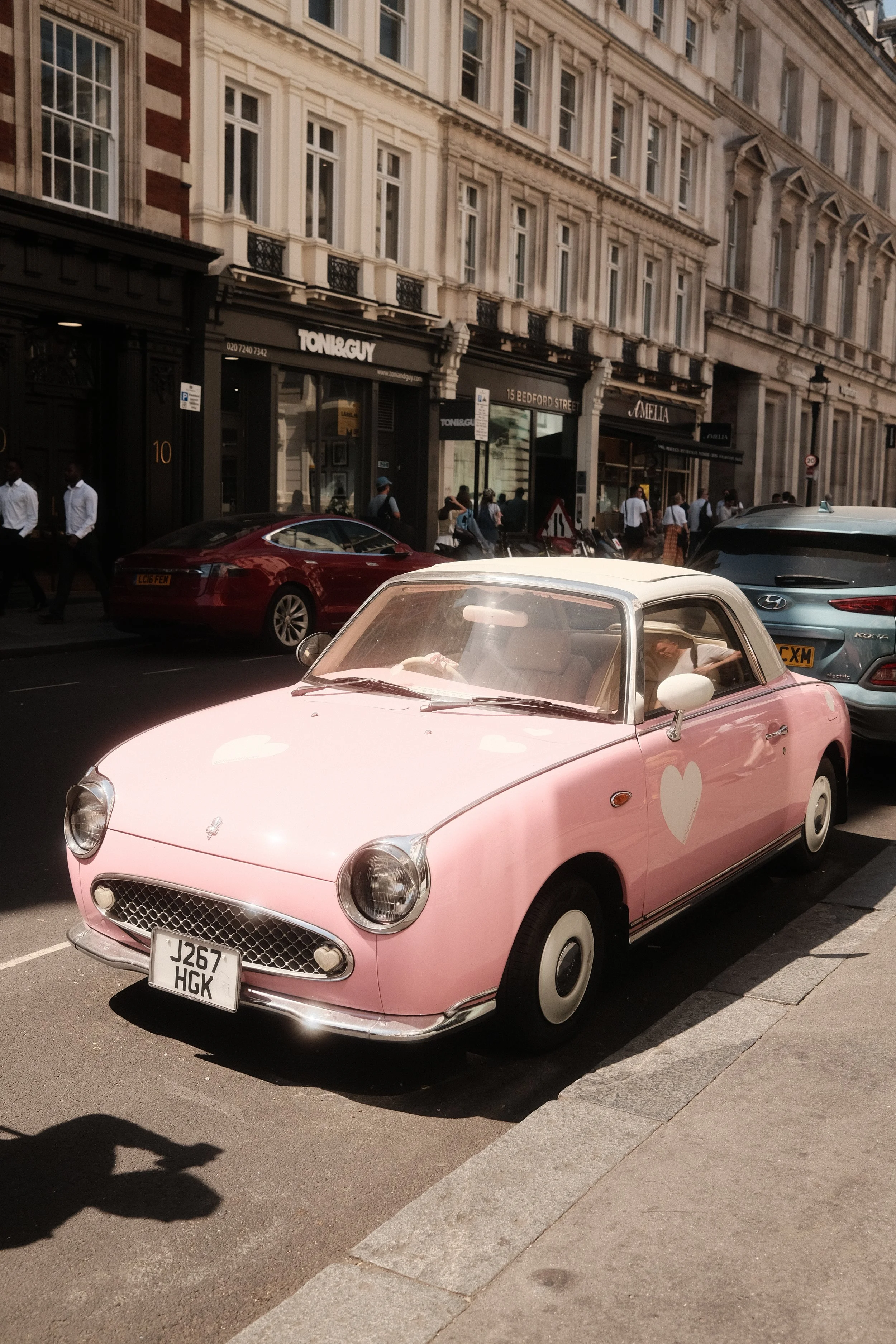 Pink vintage car with white heart on the side, parked on a street in front of a historic building with shops and pedestrians.