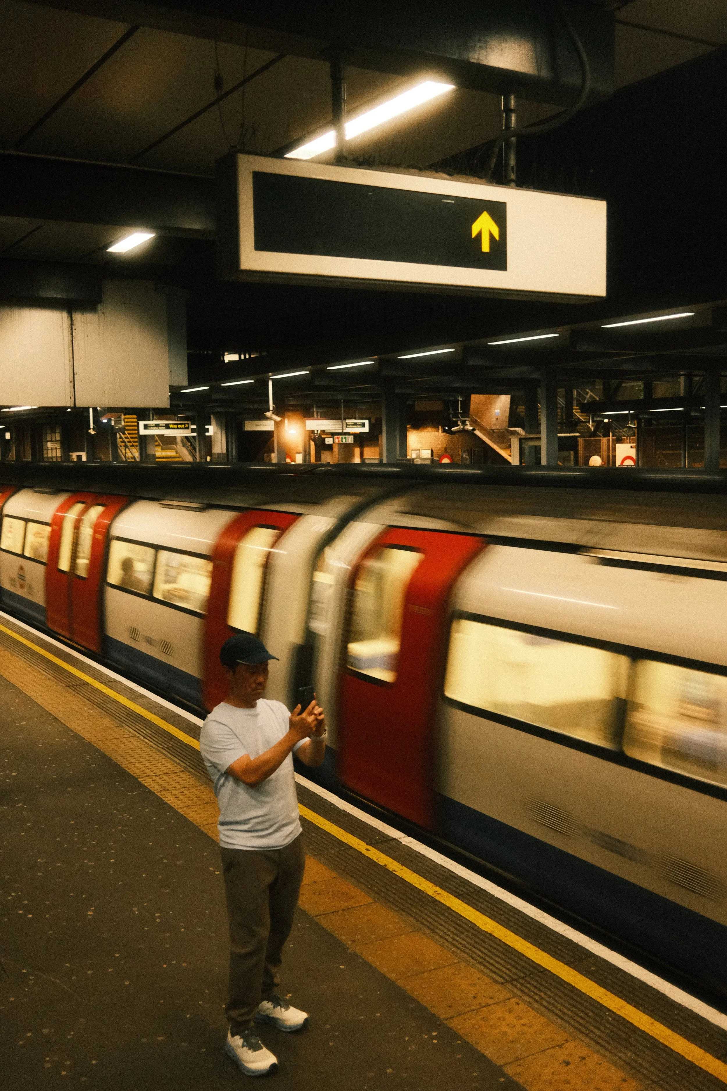 A man in a white T-shirt and dark pants stands on a subway platform, looking at his phone as a train pulls into the station behind him. There is a sign above indicating the train's direction with an upward arrow.