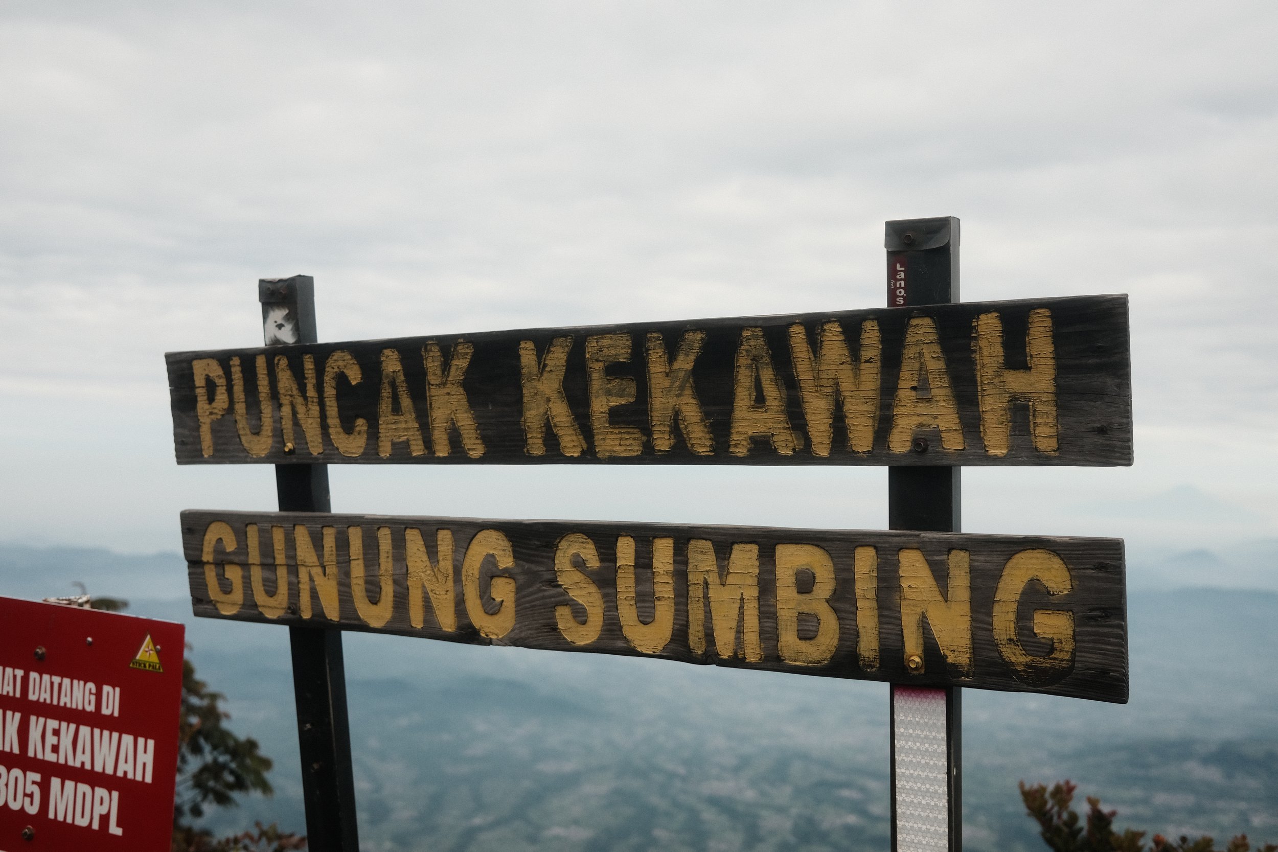 Two wooden signs with yellow text on a mountain overlook, warning about gunfire, shooting, and high-voltage power lines, with a cloudy sky in the background.