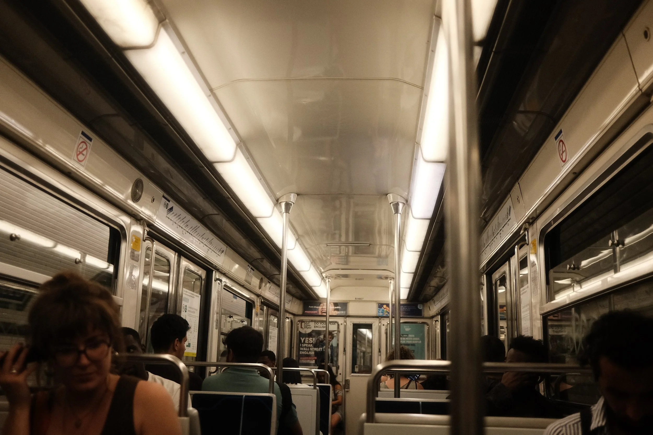 Interior of a crowded subway train with passengers seated and standing, overhead lighting, metal handrails, and window frames along the sides.