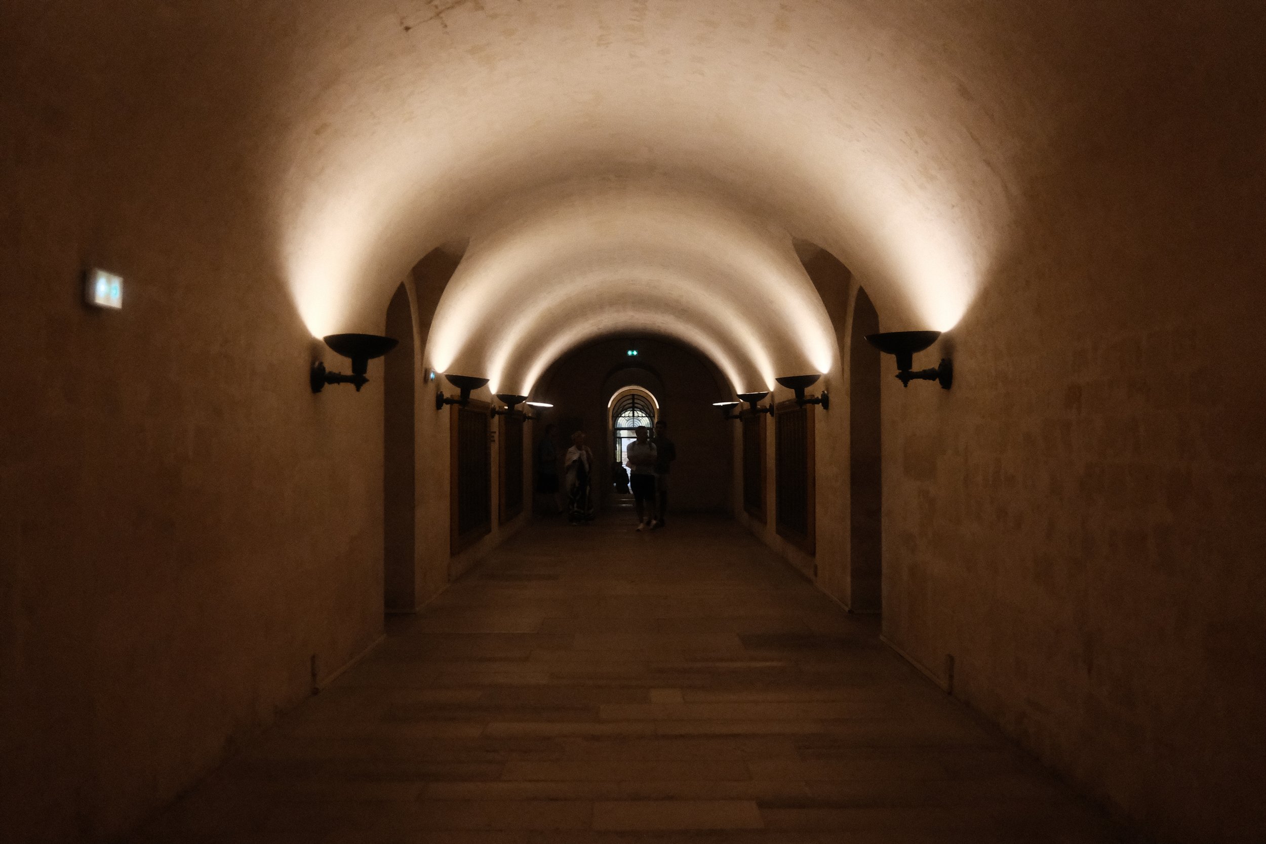 A dimly lit arched hallway with wall-mounted lamps and a wooden floor. There are a few people walking towards a window at the end of the corridor.