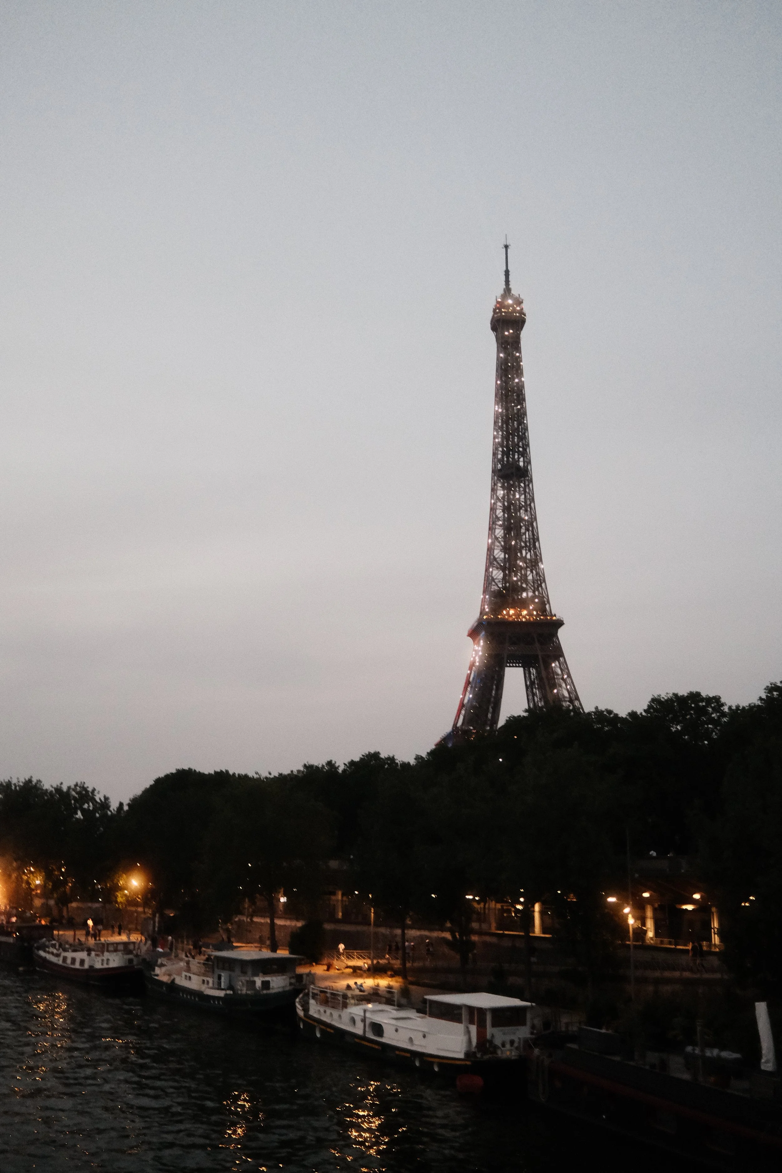 View of the Eiffel Tower illuminated at dusk, with boats docked along the river and trees in the foreground.