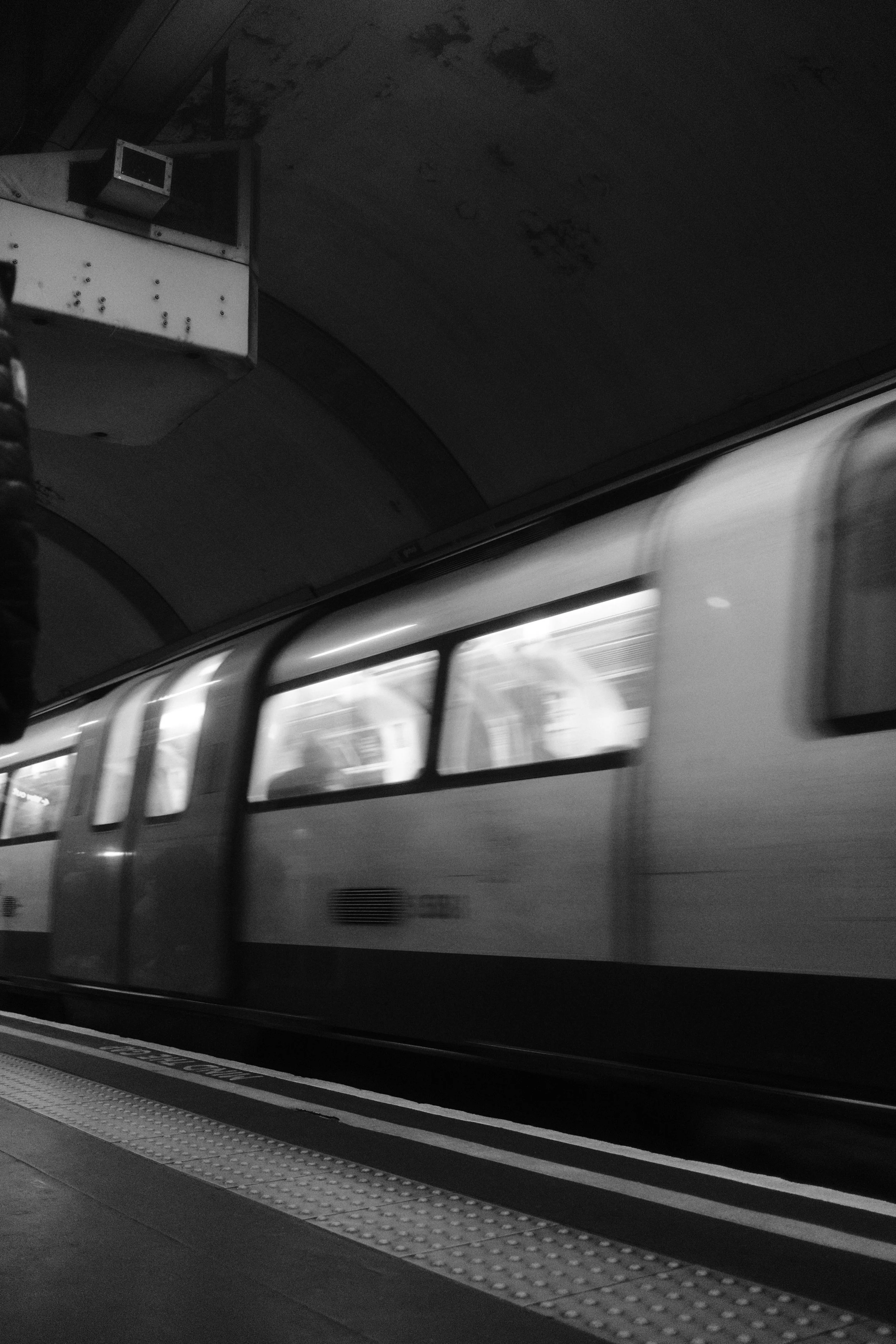 A train leaving a station at night, seen from the platform with a textured platform edge in the foreground, in a black and white photo.
