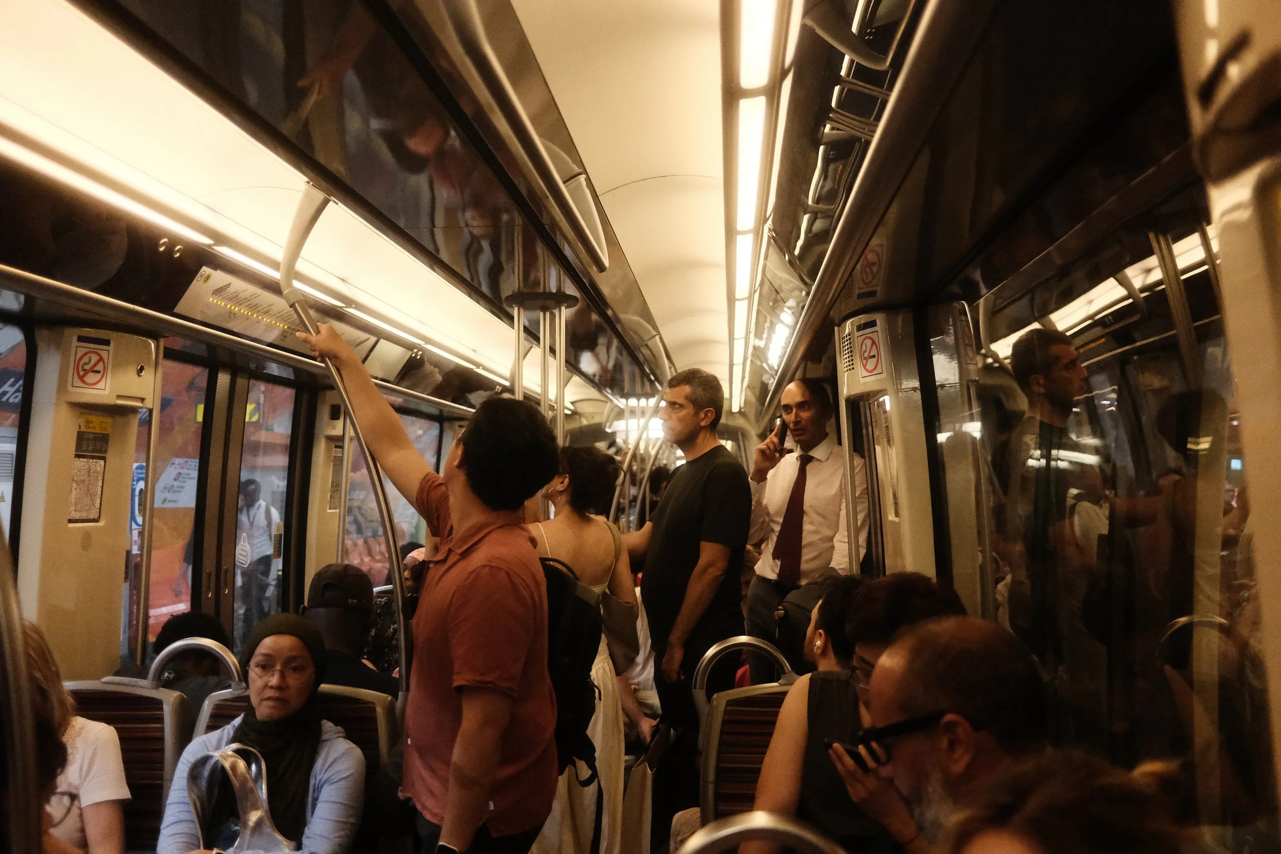 A crowded subway train with passengers standing and sitting, including a woman wearing glasses and a hijab, and individuals looking at their phones or waiting.