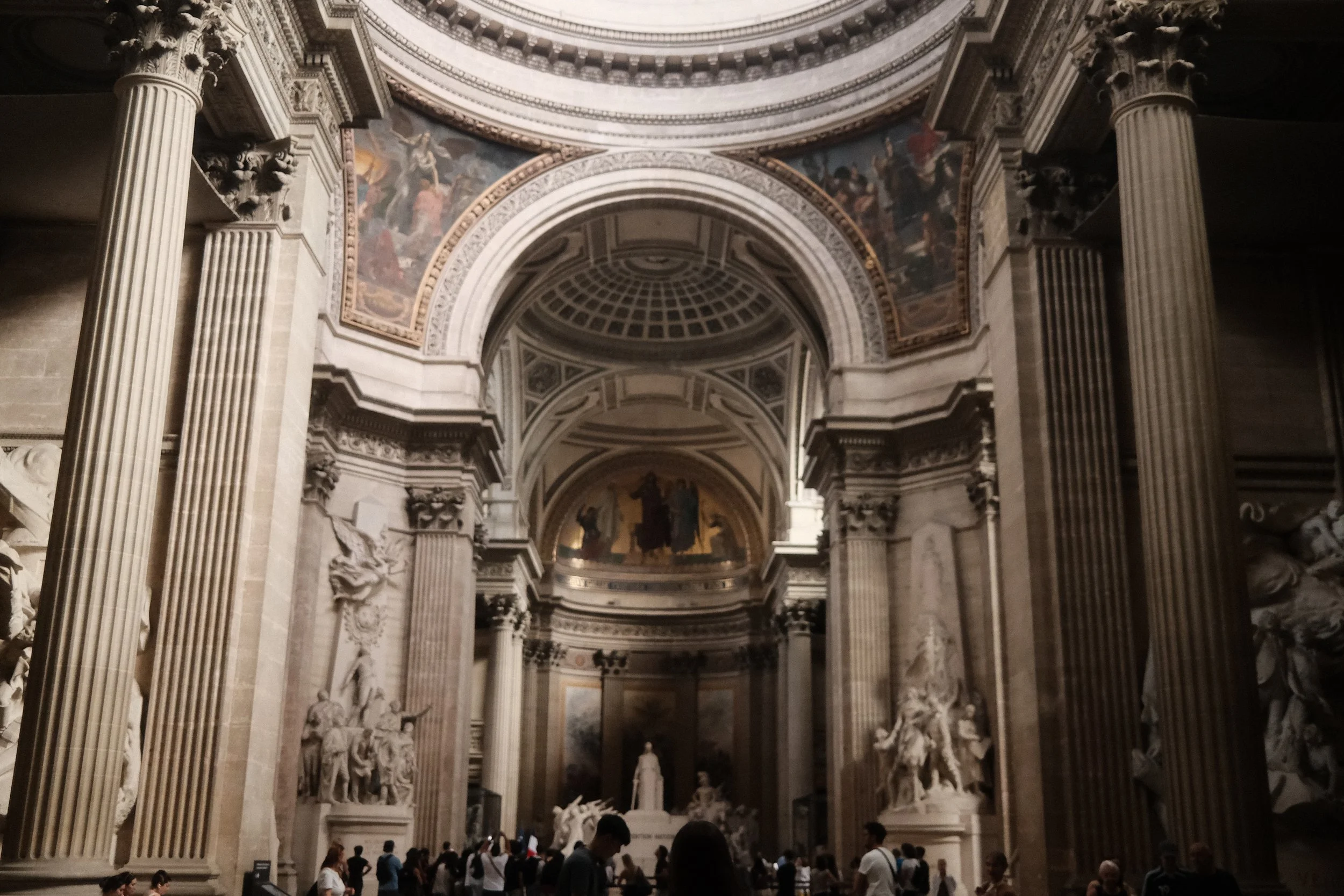 Interior of a large, ornate historic church or cathedral featuring tall, fluted columns with elaborate capitals, a decorated arch with intricate moldings, and a painted ceiling with religious artwork and frescoes. There are visitors or tourists stand