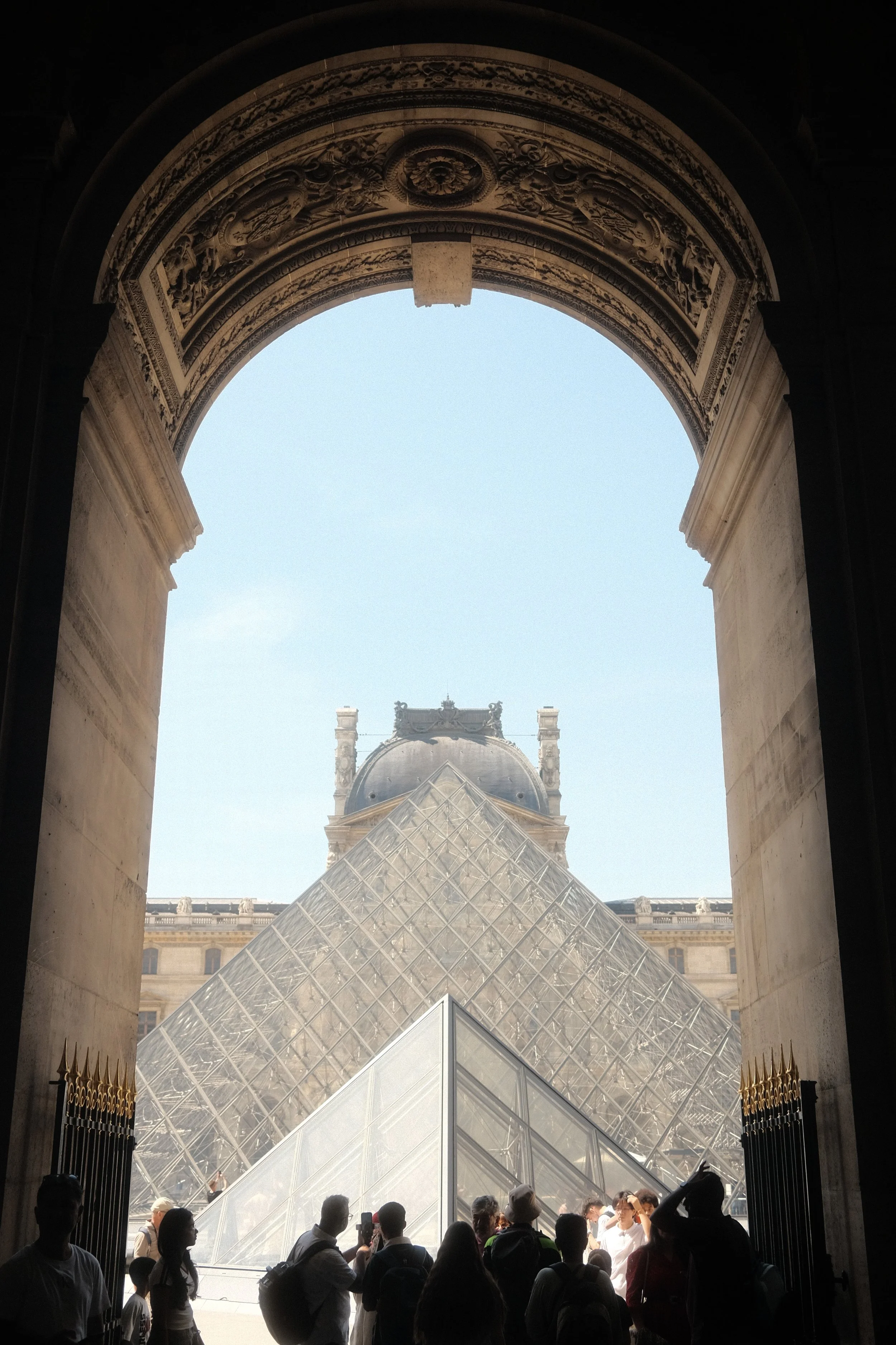 View of the Louvre Pyramid through an archway at the Louvre Museum in Paris, with visitors in the foreground.