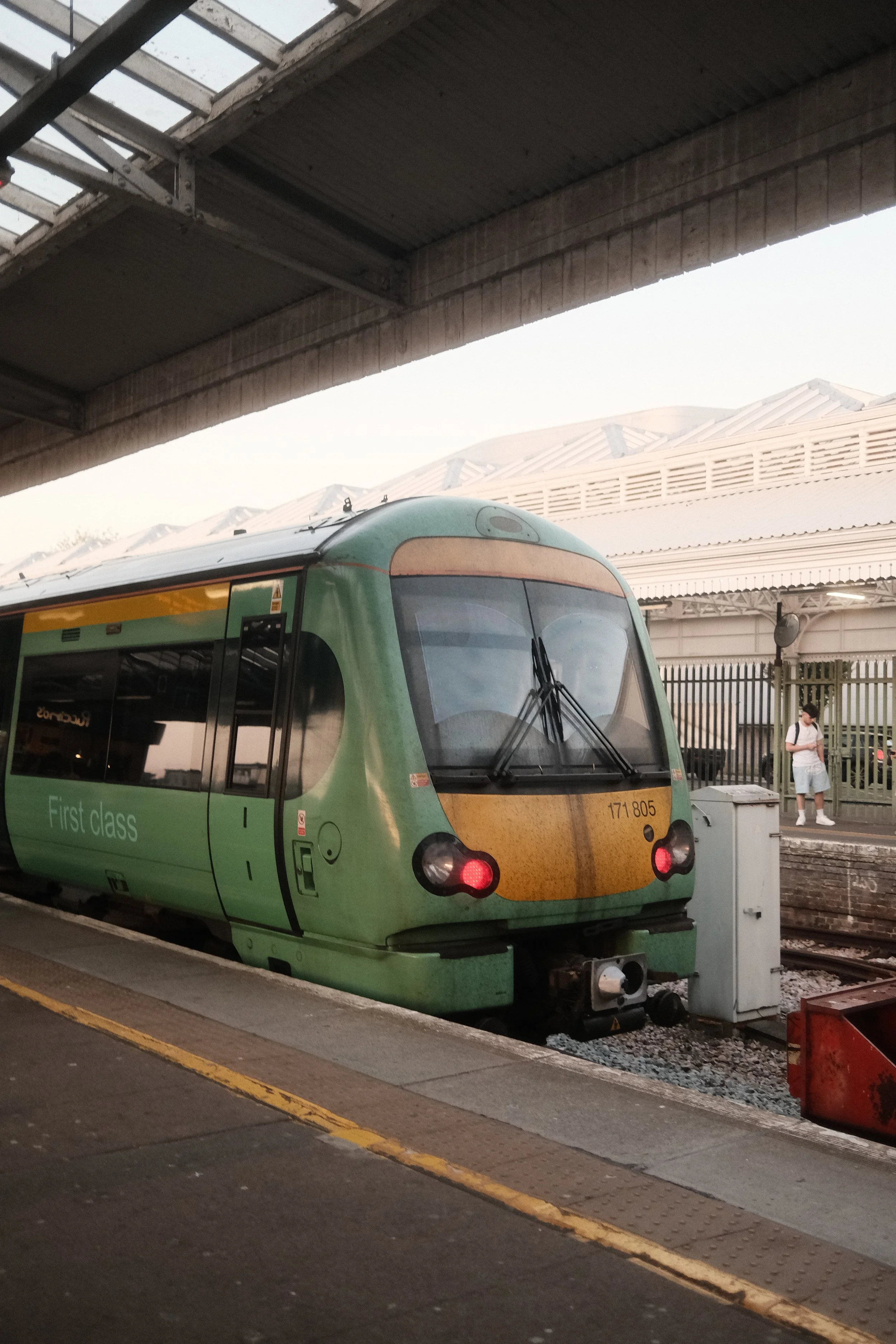 A modern green train with a yellow front at a station platform, with a person waiting nearby.