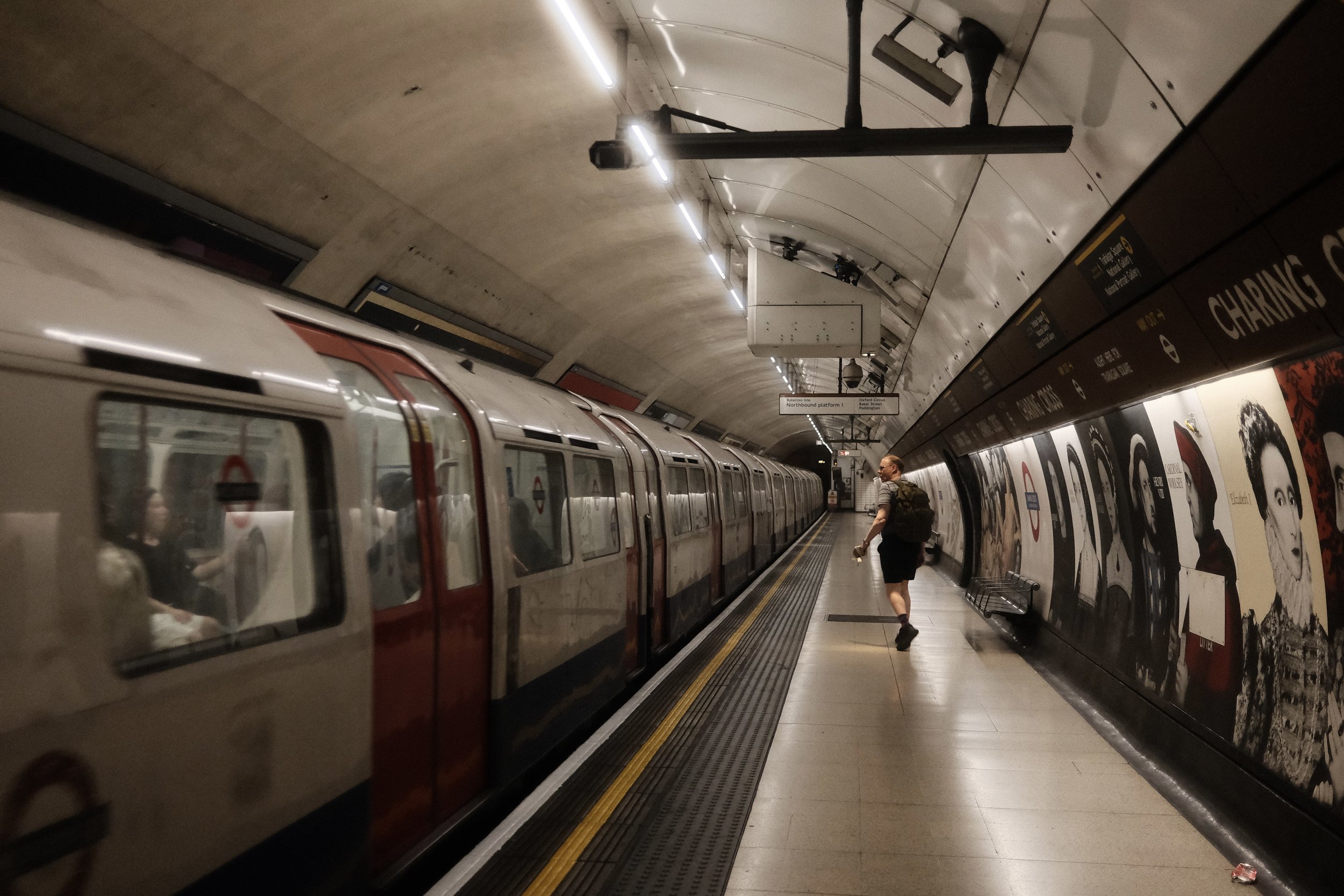 Underground metro station with train on the platform, a person walking beside the train, and advertisements on the wall. Sign overhead indicates 'Northbound platform 1' at a London Tube station.