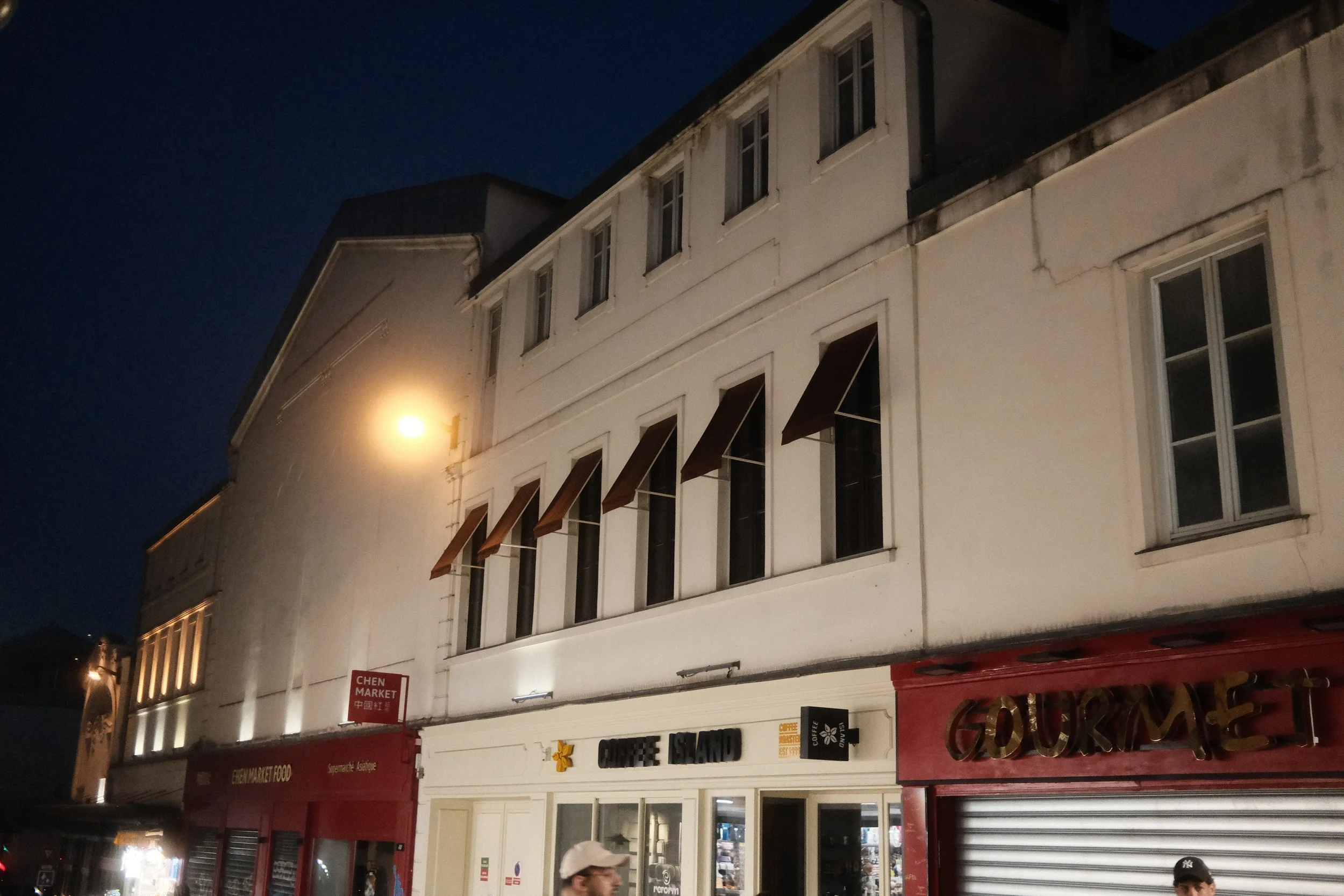 Nighttime street scene featuring a multi-story building with a white facade, illuminated shop signs, and people walking on the sidewalk.
