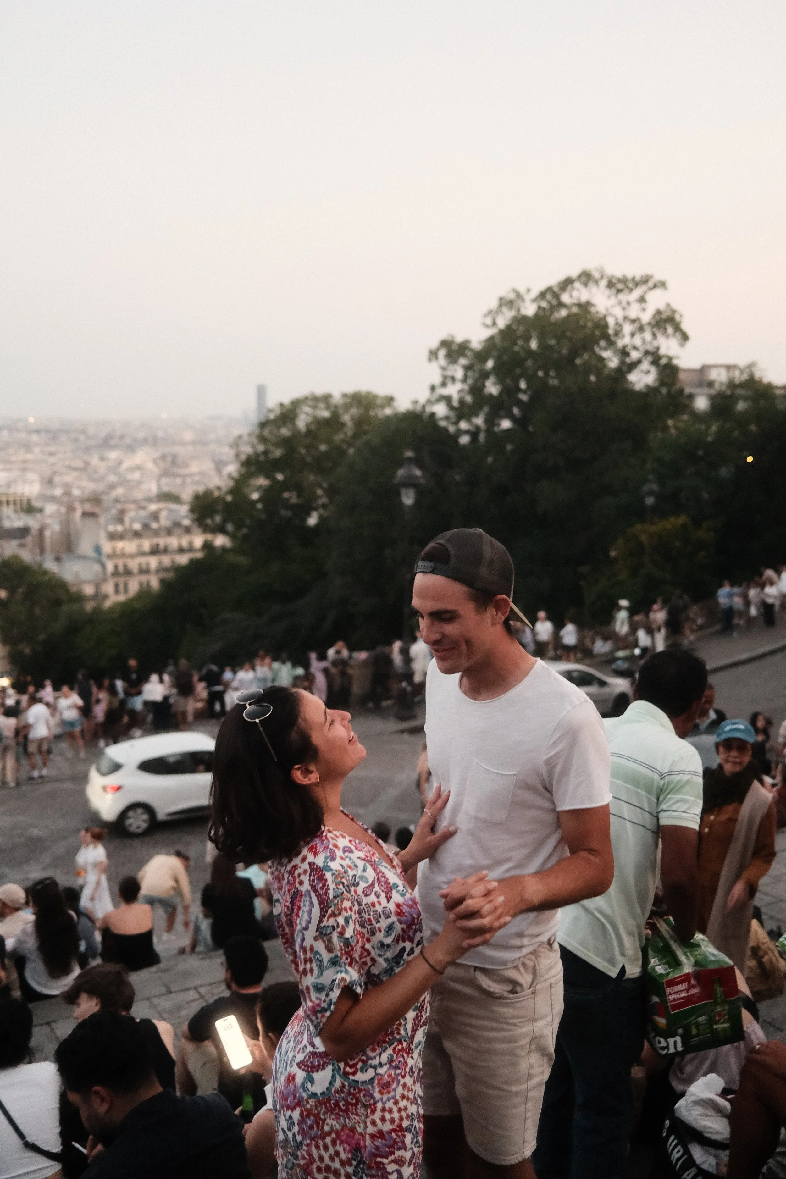 A man and a woman are dancing closely together amidst a crowd on an outdoor staircase. The man is tall, wearing a white t-shirt and beige shorts, smiling while looking down at the woman. The woman has dark hair, wearing a colorful printed dress, smil