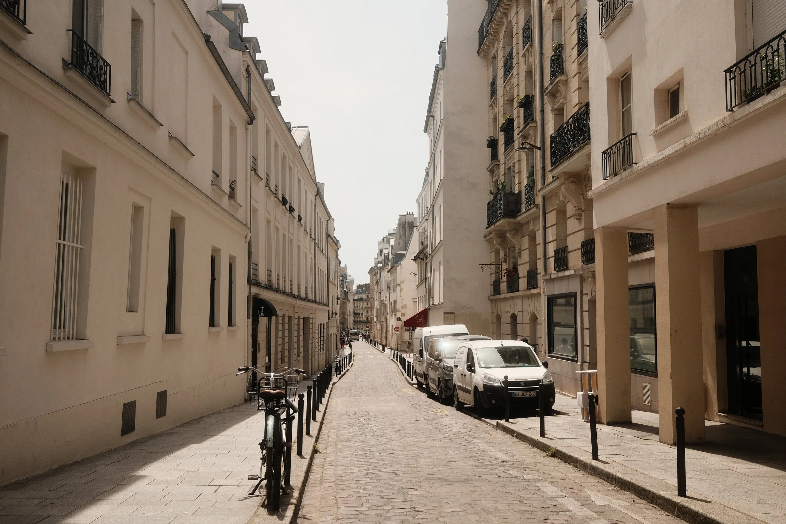 A narrow cobblestone street in a city, lined with cream-colored buildings with wrought-iron balconies. Parked cars and bicycles are on the street, with a few pedestrians visible.