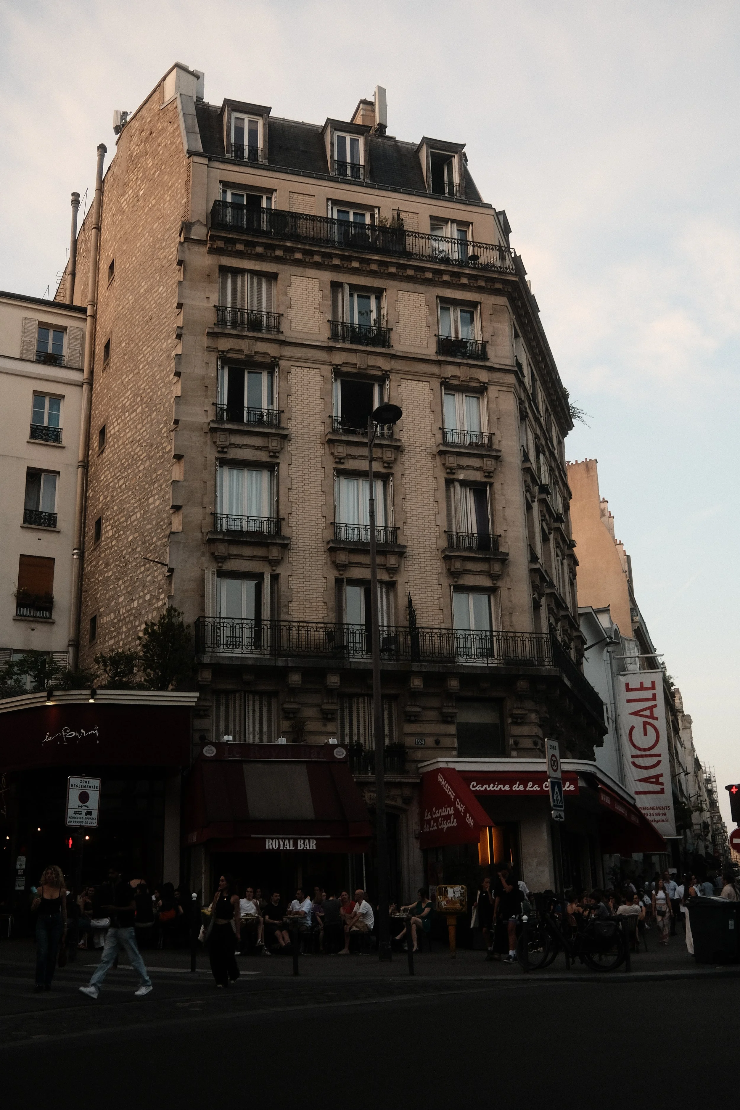 Parisian street scene in the evening with a multi-story building, outdoor café with people sitting, and pedestrians walking.