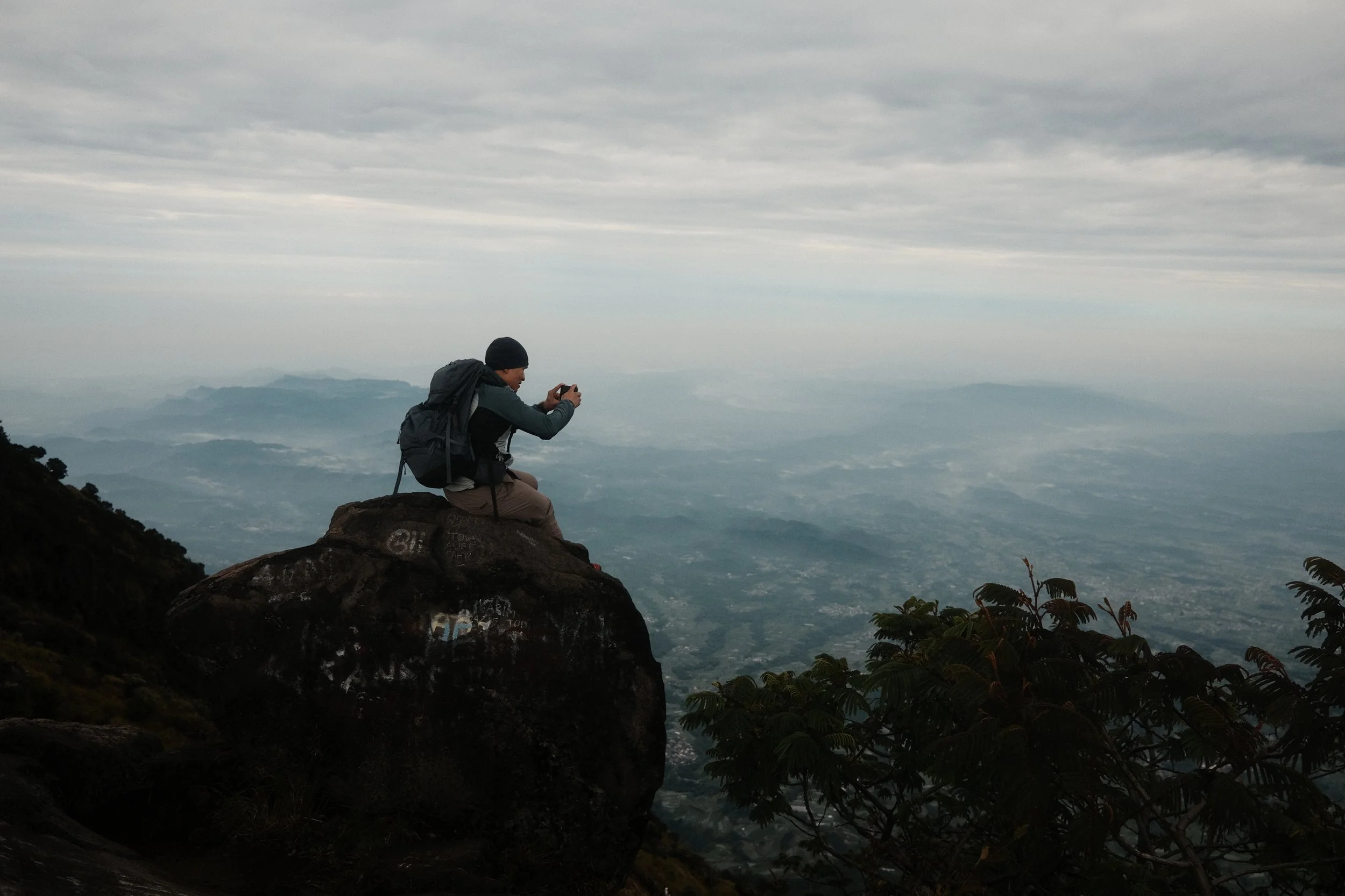A person sitting on a large rock on a mountain peak, wearing a backpack and a beanie, taking a photo with a camera as clouds and mountains are visible in the distance.