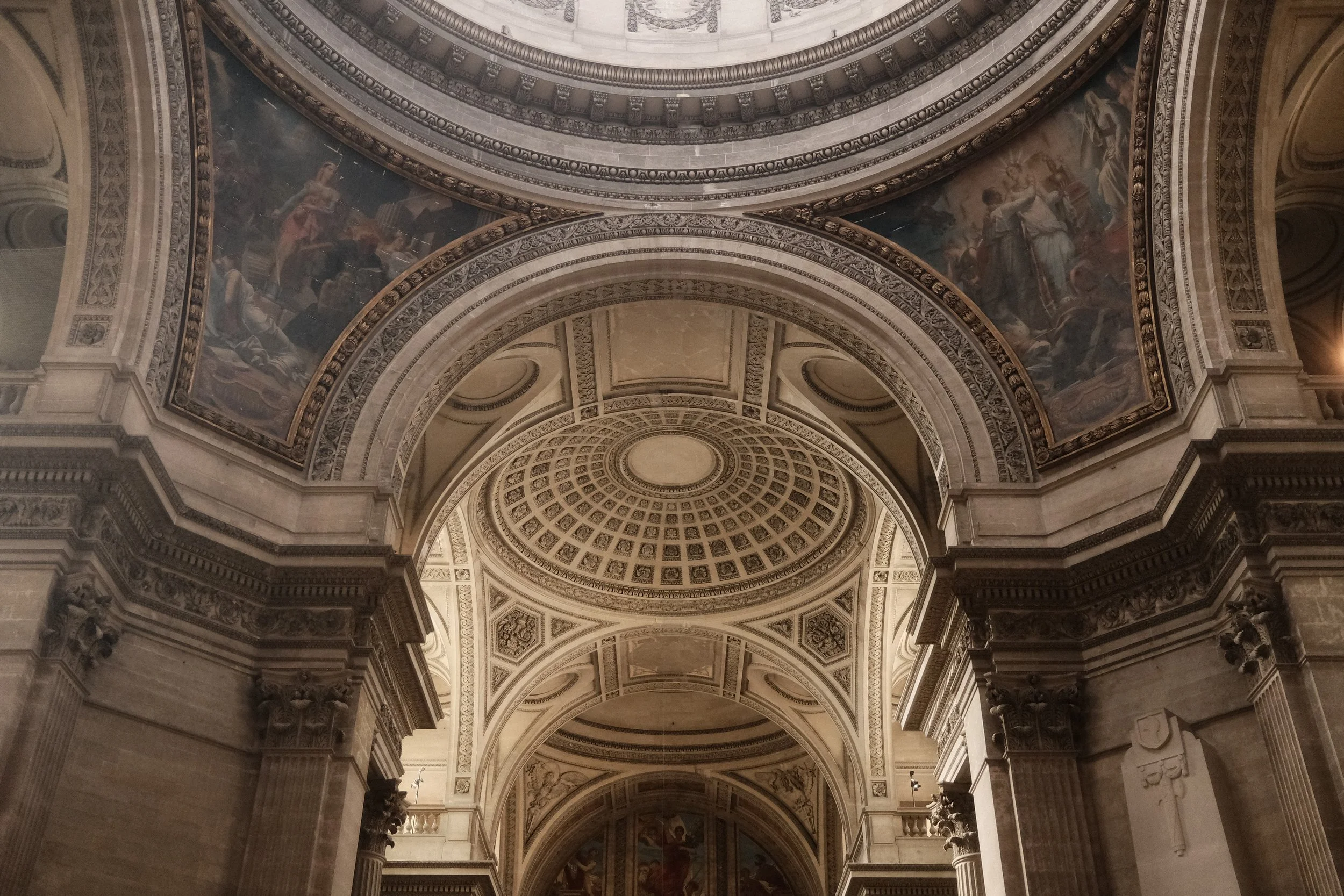 The interior of a grand, ornate building with high vaulted ceilings decorated with intricate patterns and paintings, supported by large columns.