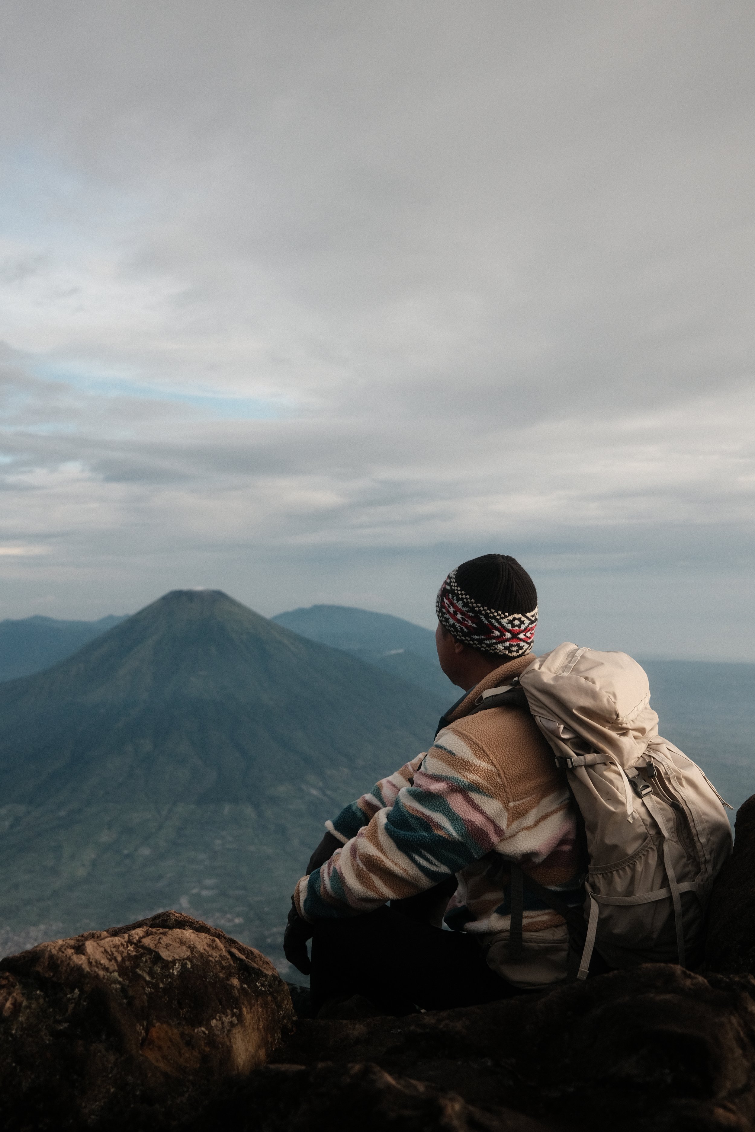 A person wearing a striped sweater, beige backpack, and a knitted hat sitting on rocks overlooking a mountain landscape with a volcano in the distance under a cloudy sky.