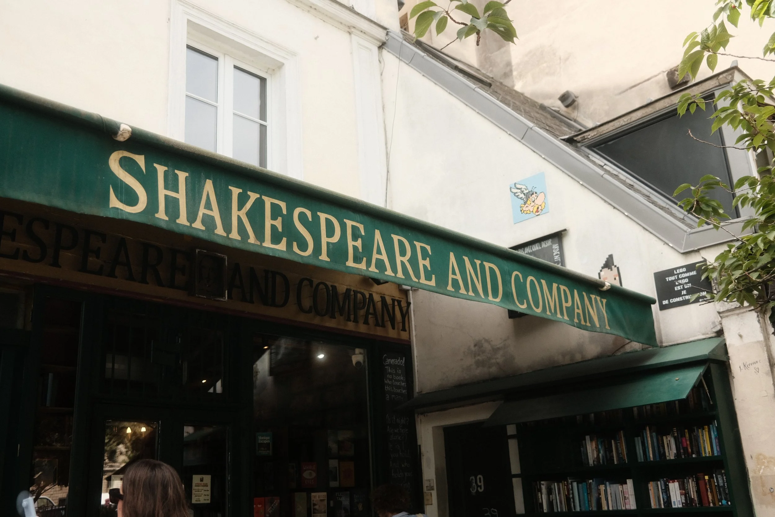 Storefront with green awning displaying the text 'SHAKESPEARE AND COMPANY' and shelves of books inside, with a small picture of a winged pig on the wall and a person with long hair and sunglasses standing outside.