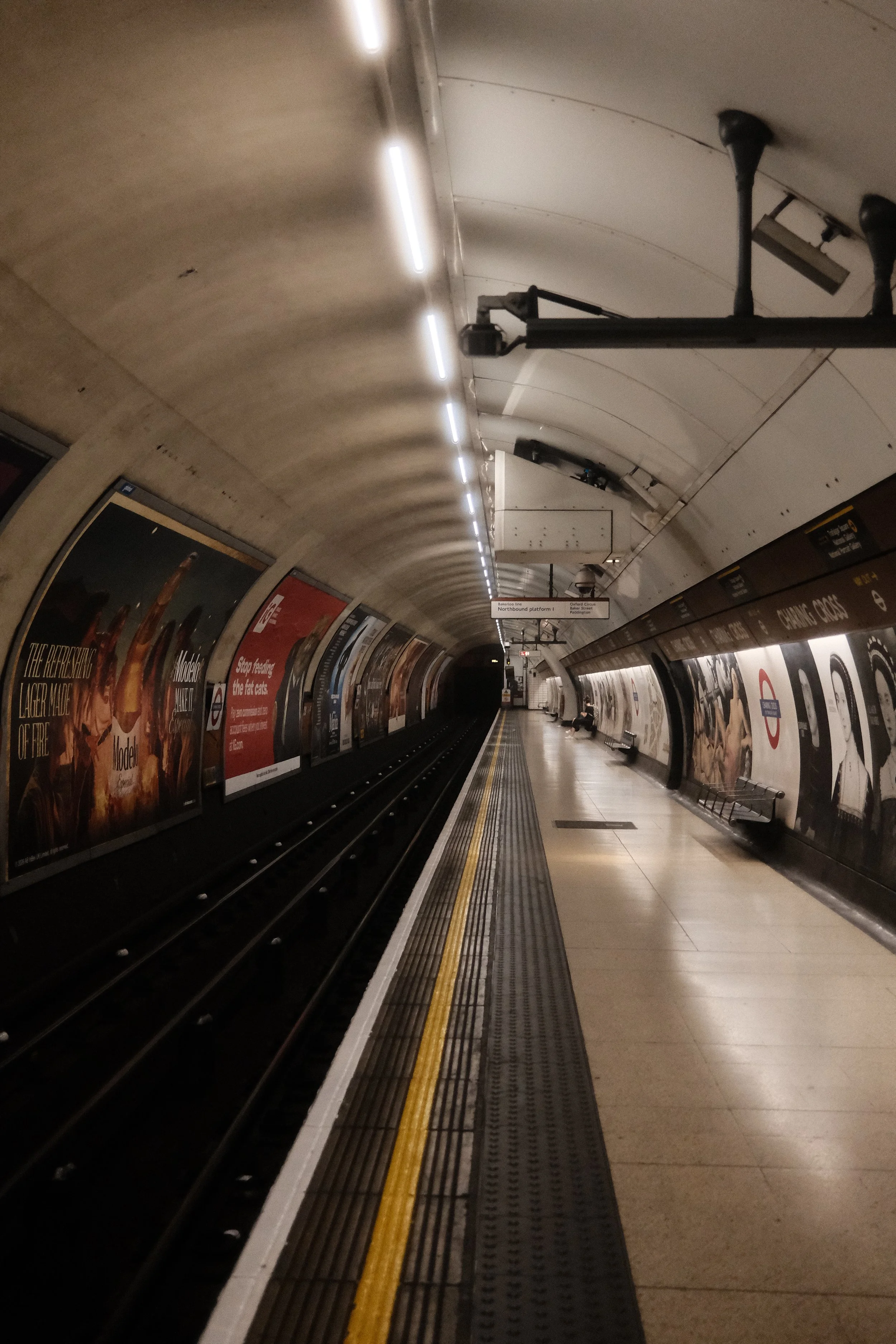An empty underground subway station platform with advertisements on the walls, a digital sign hanging from the ceiling, and a bench on the right side.