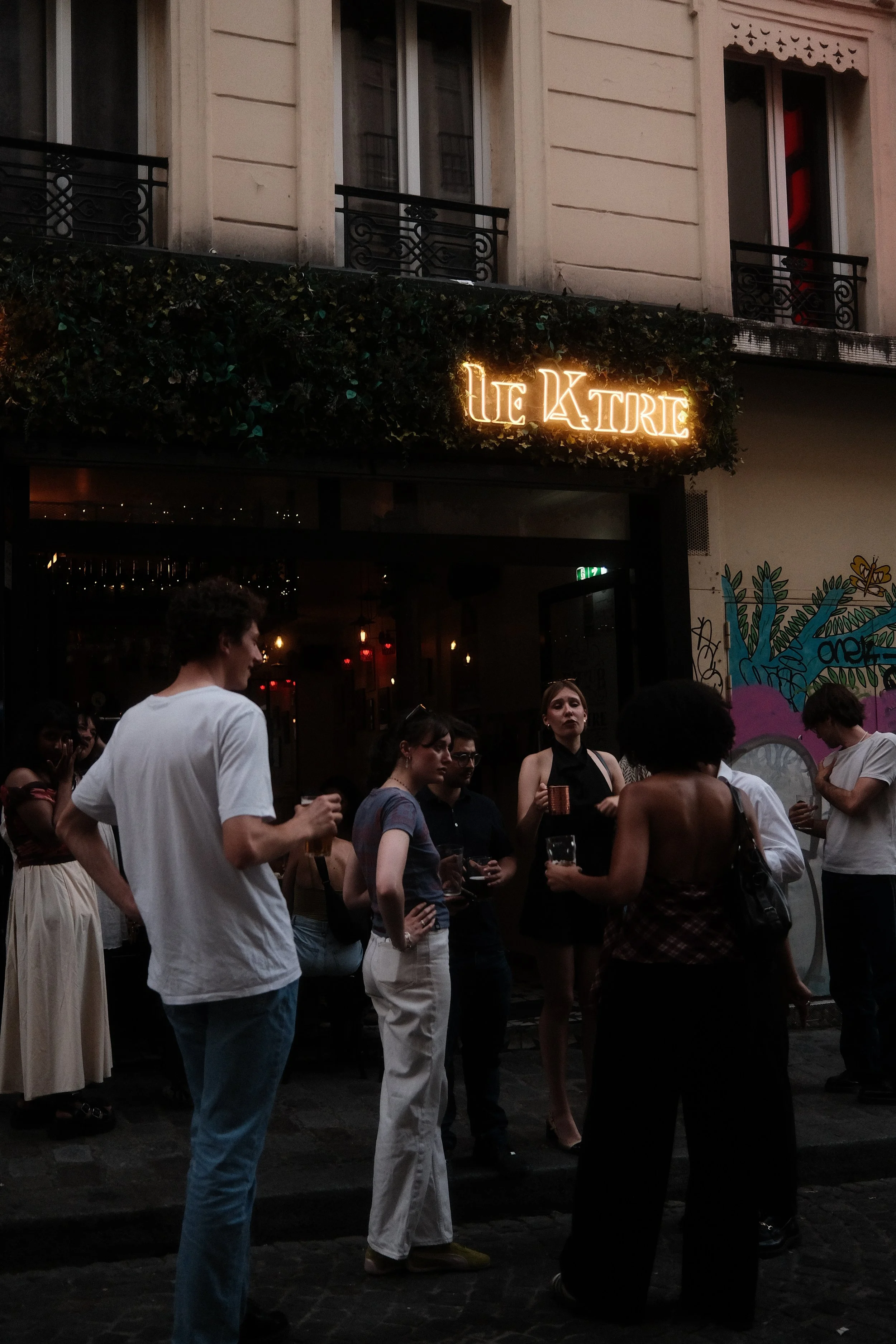 People gathered outside a venue named 'Le Katre' with a neon sign on the building. The scene appears to be in the evening, with some individuals engaged in conversation, holding drinks, and socializing.
