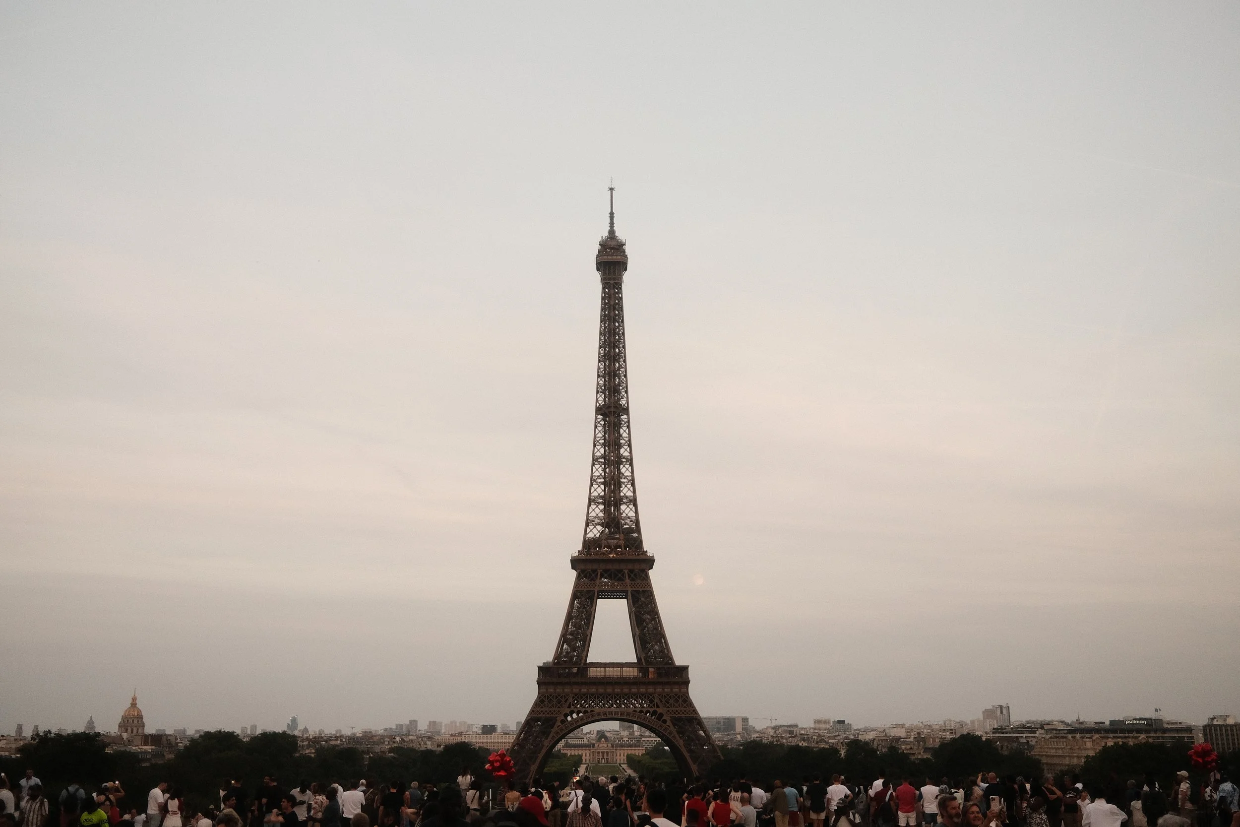 Eiffel Tower with a large crowd of people at its base, Paris skyline in the background, overcast sky.