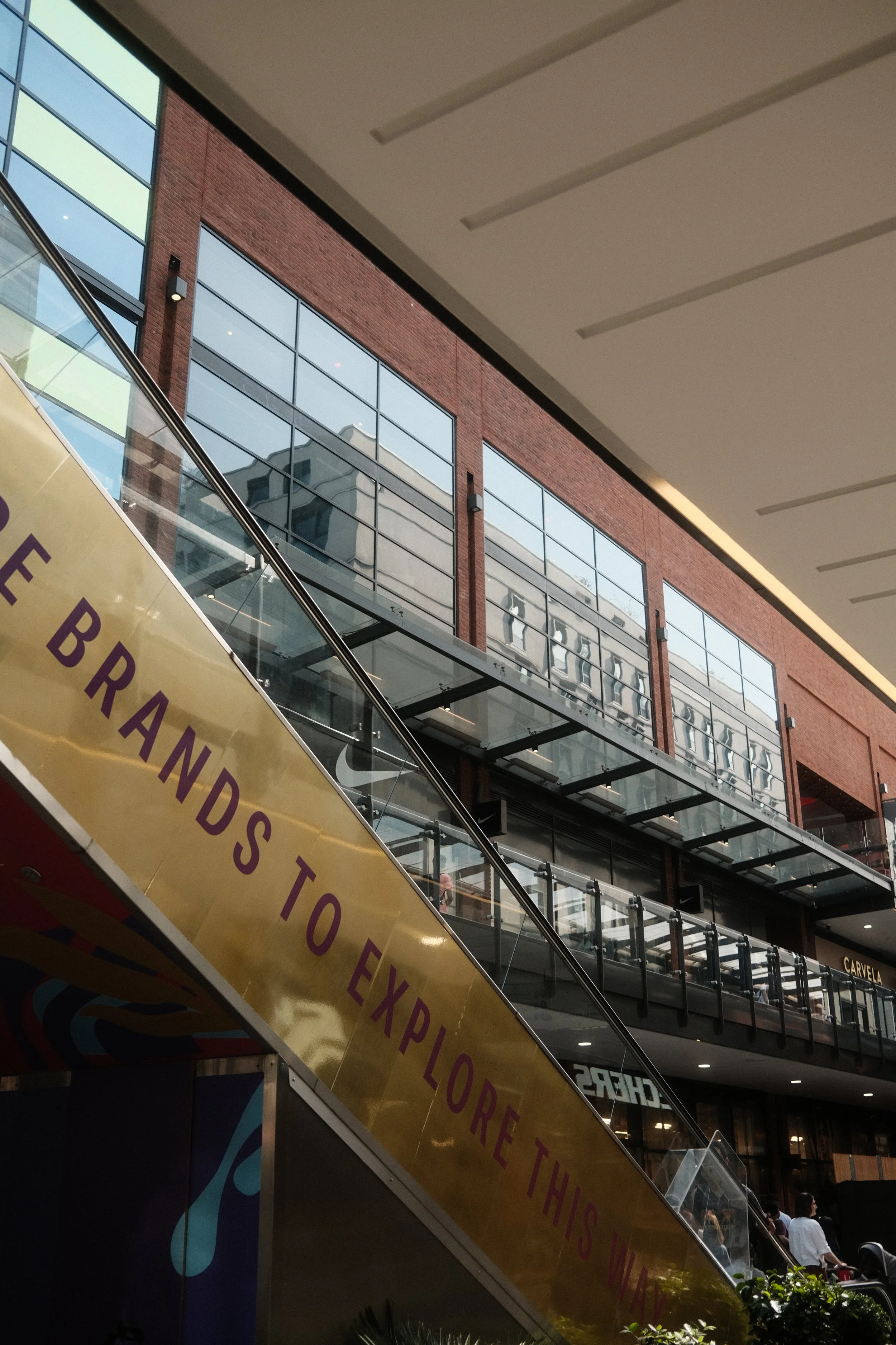 Interior view of a modern shopping mall with large glass windows, an escalator, and a yellow sign that reads 'BRANDSTO' and part of another word. People are visible walking in the background.