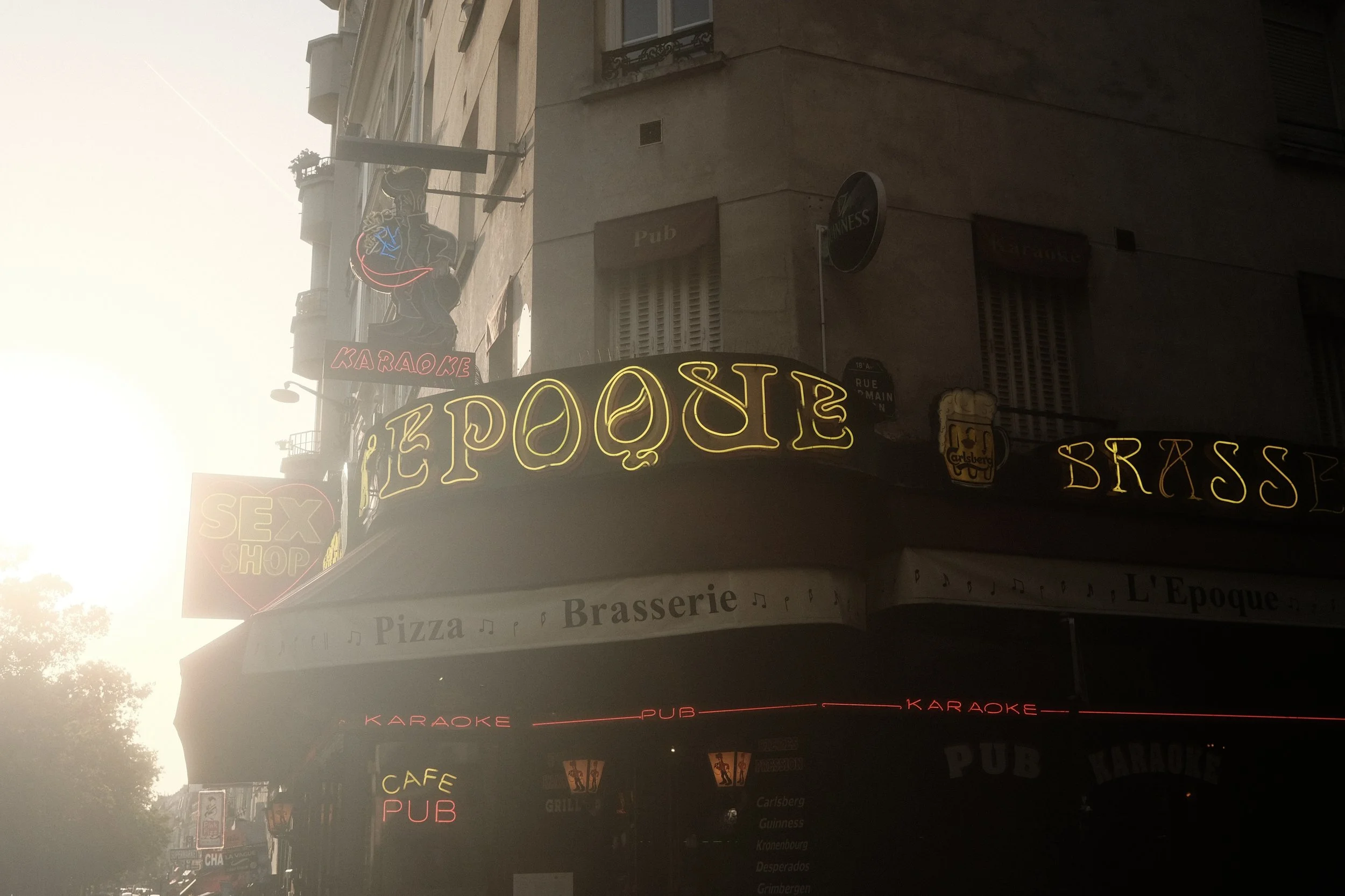 A street view with multiple illuminated signs on a building, including signs for a pub, karaoke, and a sex shop. The signs feature neon lighting in various colors, including yellow, red, and blue. The building appears to be in an urban area with othe