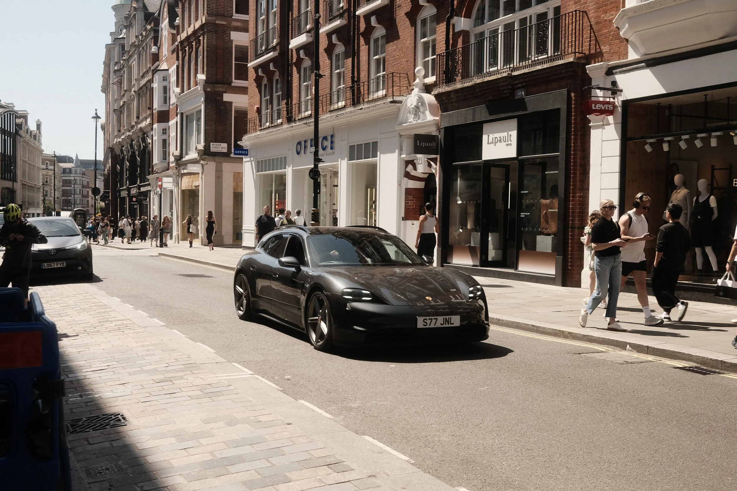 Street scene with brick buildings, shops, pedestrians walking on the sidewalk, and two parked cars, including a black sports car in the foreground.