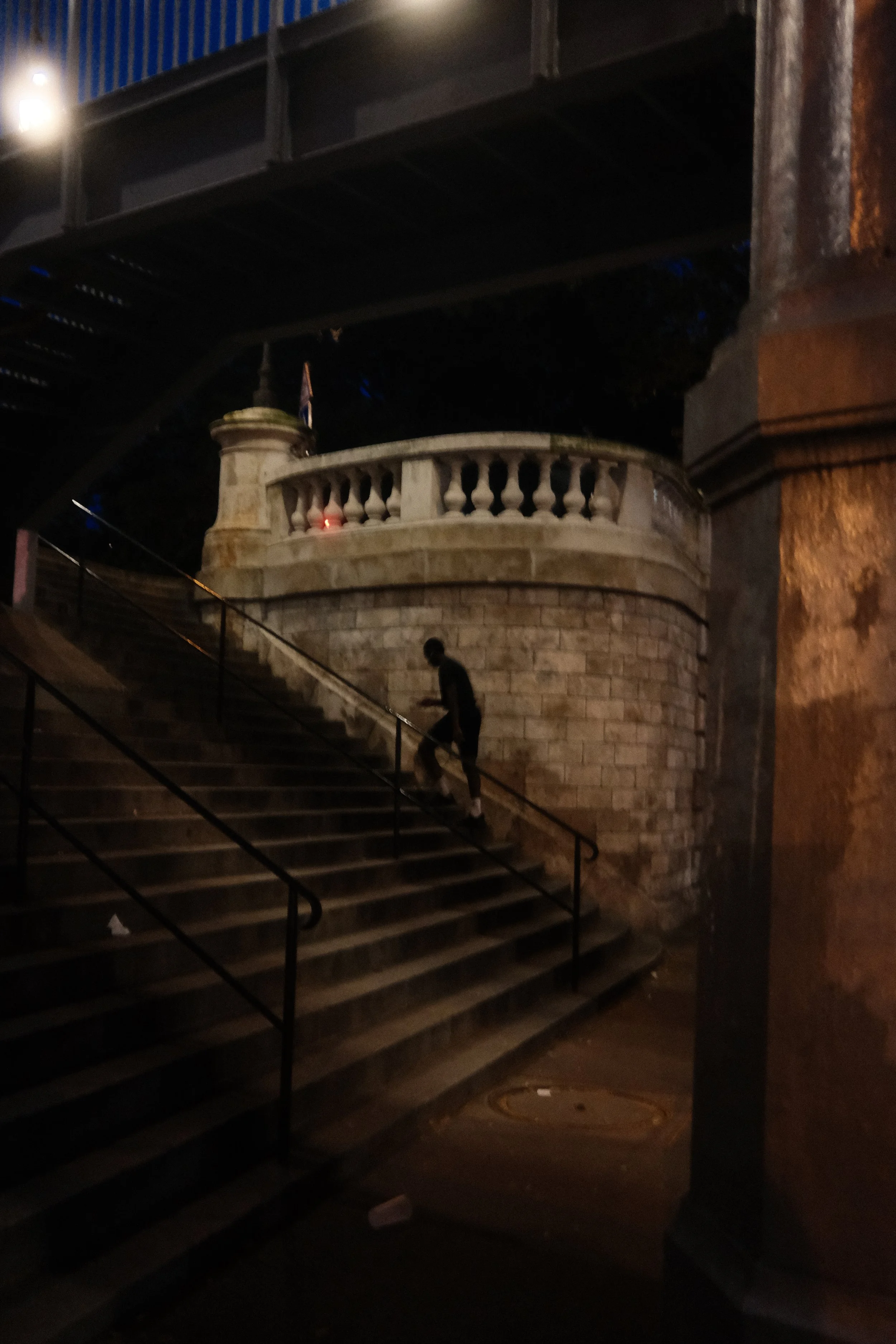 Nighttime scene of a city street with stairs leading up to a stone balcony, a person walking down the stairs, and a streetlamp illuminating the area.