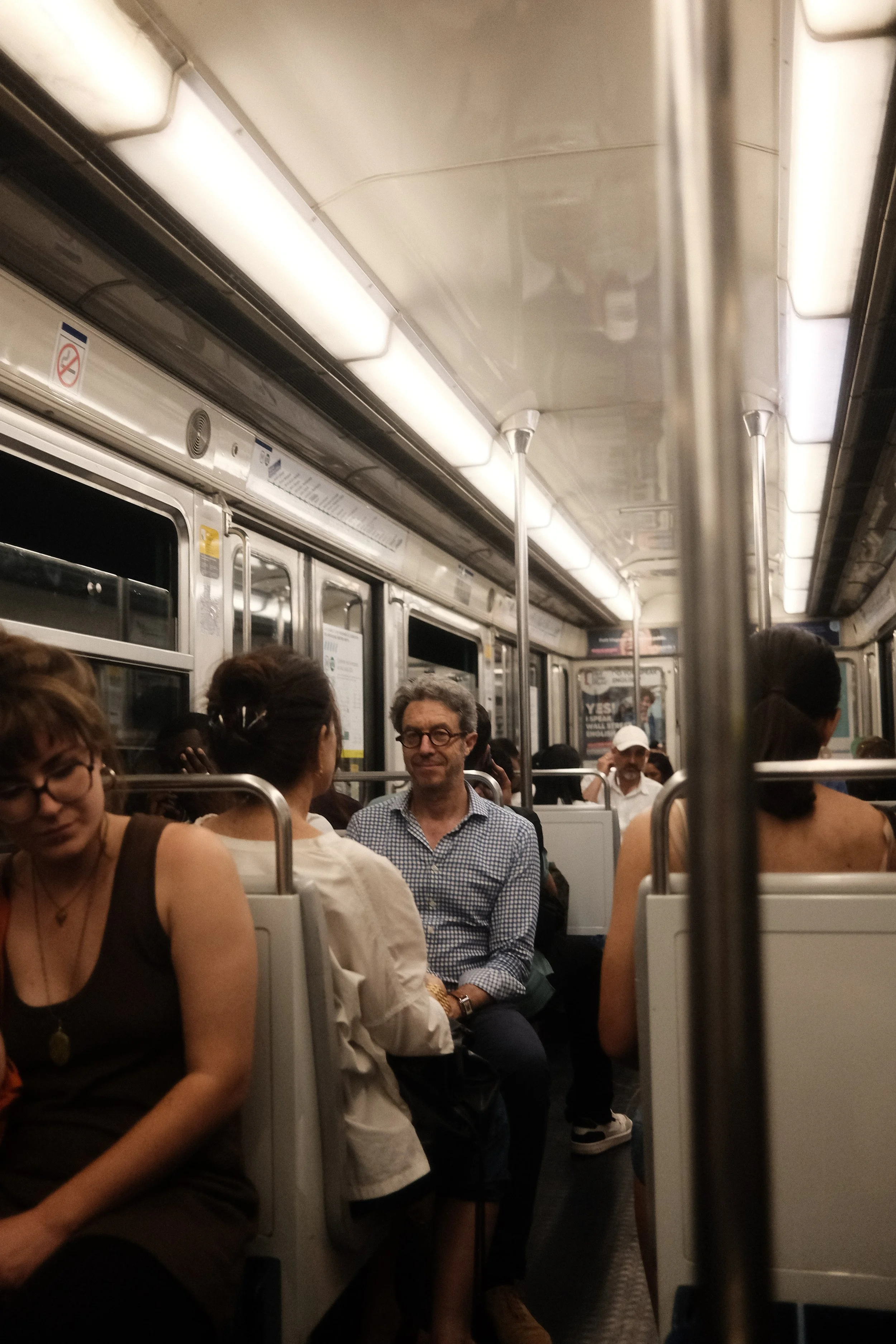 People sitting closely inside a train or subway car, with overhead lights and metal poles for standing passengers.