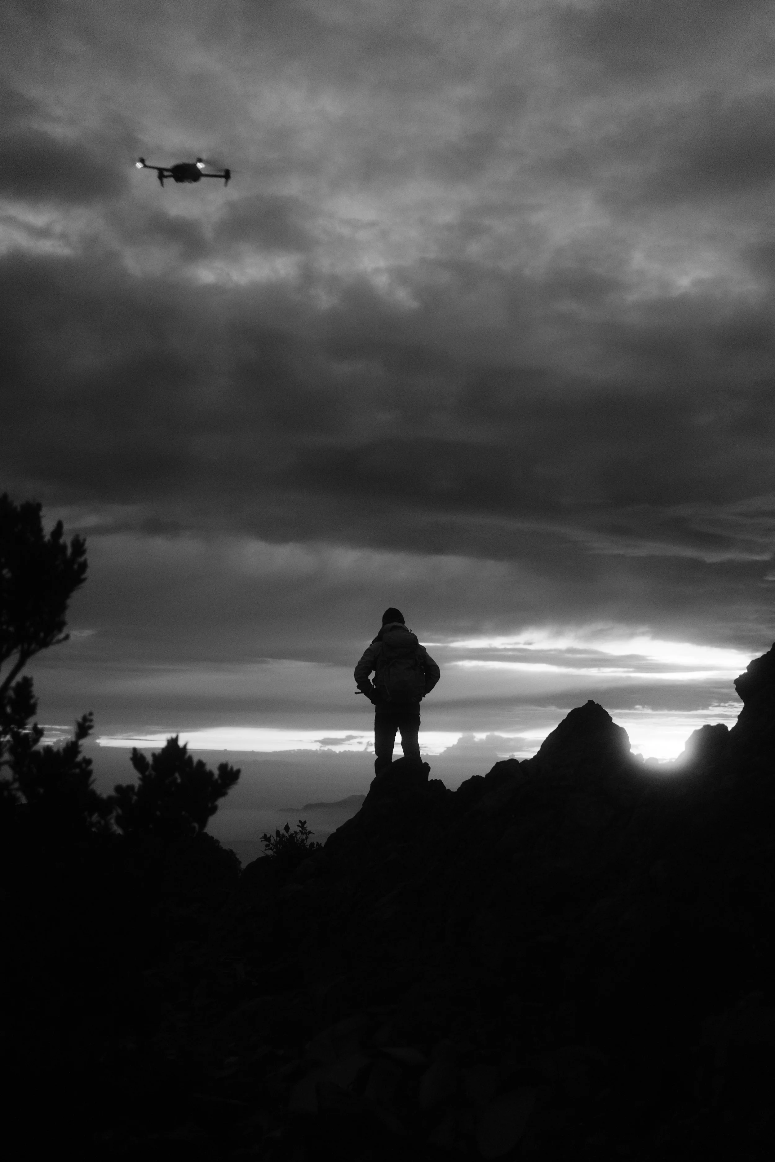 A person standing on a rocky hilltop during dusk with cloudy skies and a drone flying overhead.