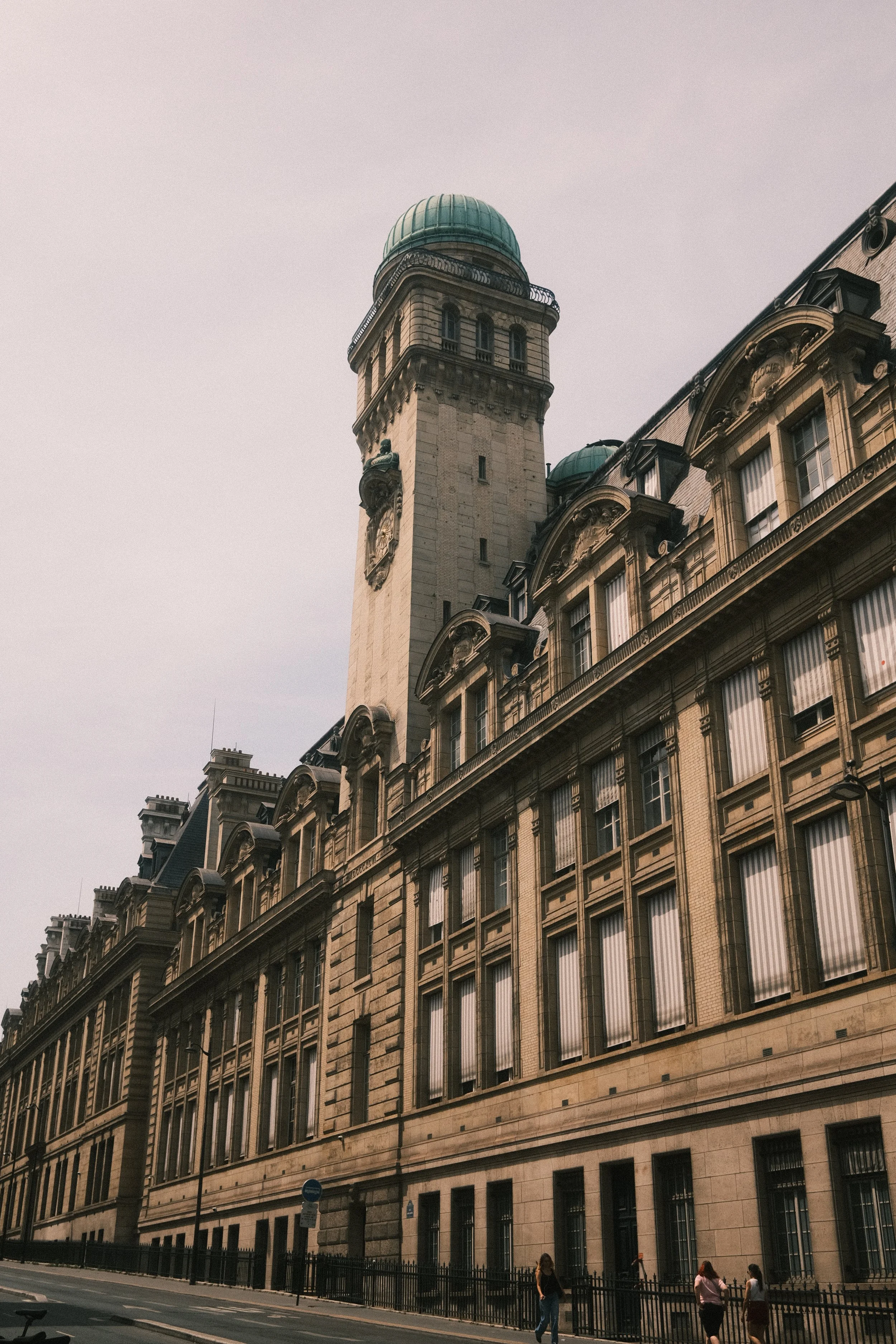 A historic building with a clock tower and green-domed roof, featuring classical architectural details and multiple windows, located on a city street with pedestrians.