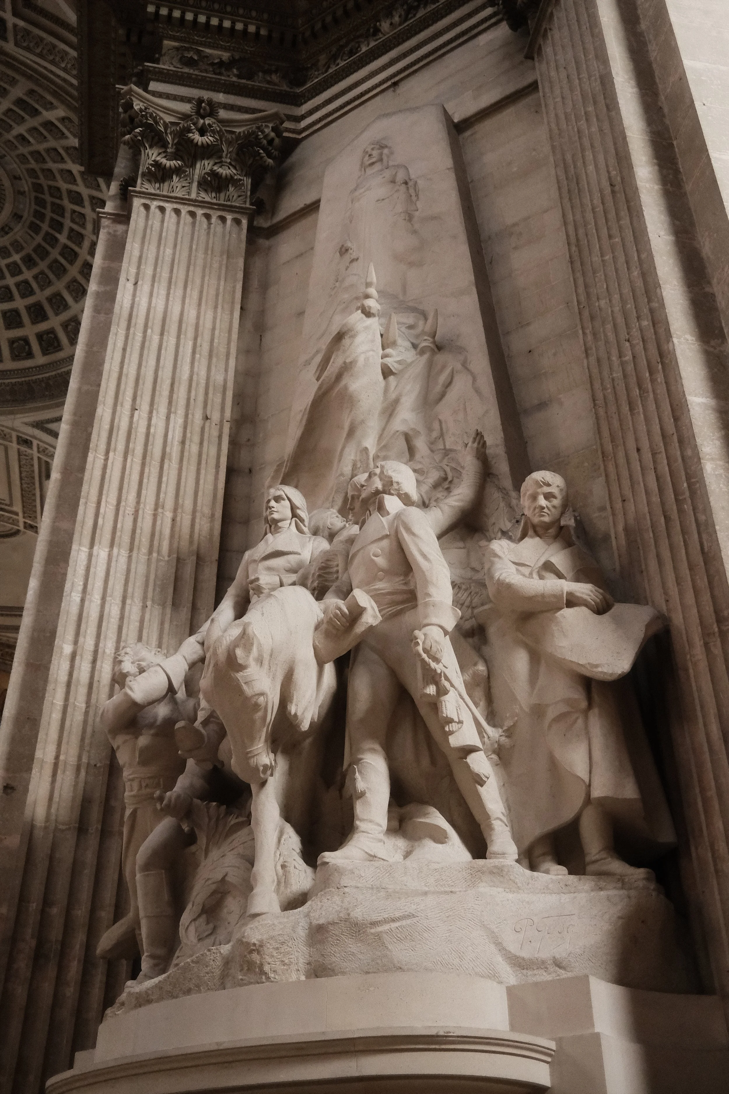 A marble sculpture inside a grand building with tall columns and detailed ceilings. The sculpture depicts a group of historical figures, some on horseback, with one figure on a horse in the foreground holding a flag, and others standing or sitting ar