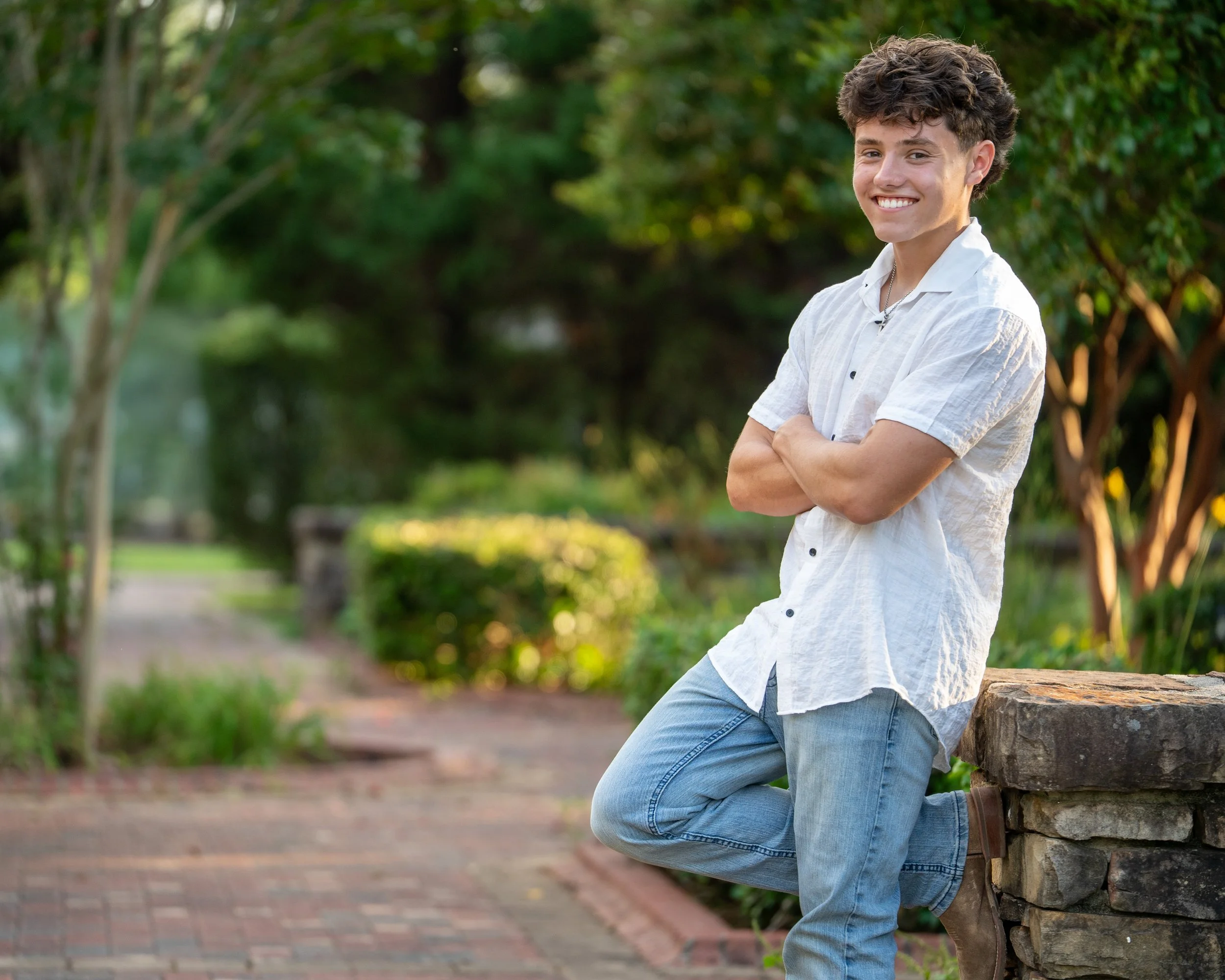 A young man in a white top and blue jeans poses against green foliage leaning against a brick wall at E. Carroll Joyner Park, Wake Forest, North Carolina for High School Senior Photos