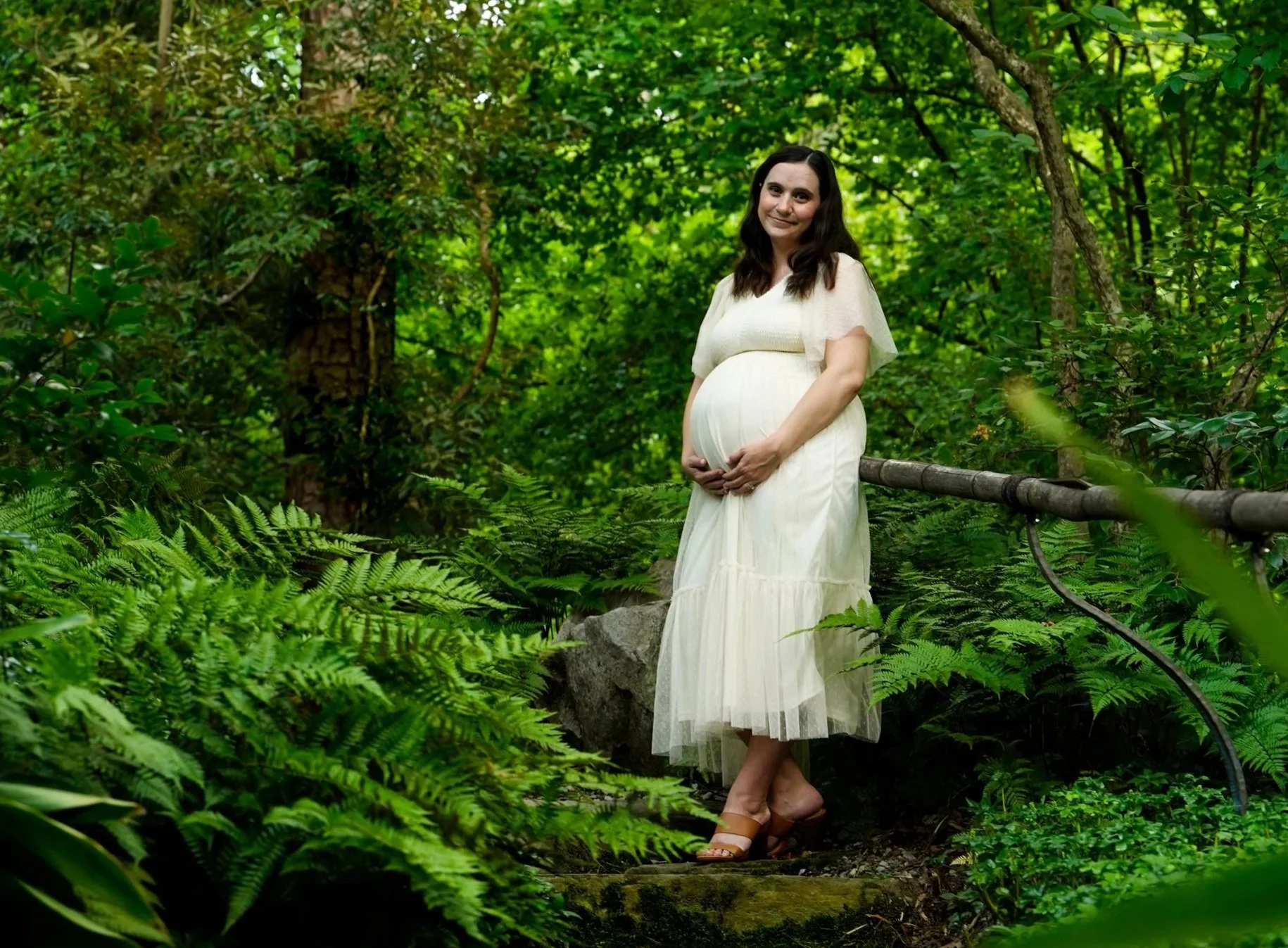 A pregnant woman in a white dress standing on a forest trail, holding her baby belly, surrounded by lush green foliage at Duke Gardens, Durham North Carolina.