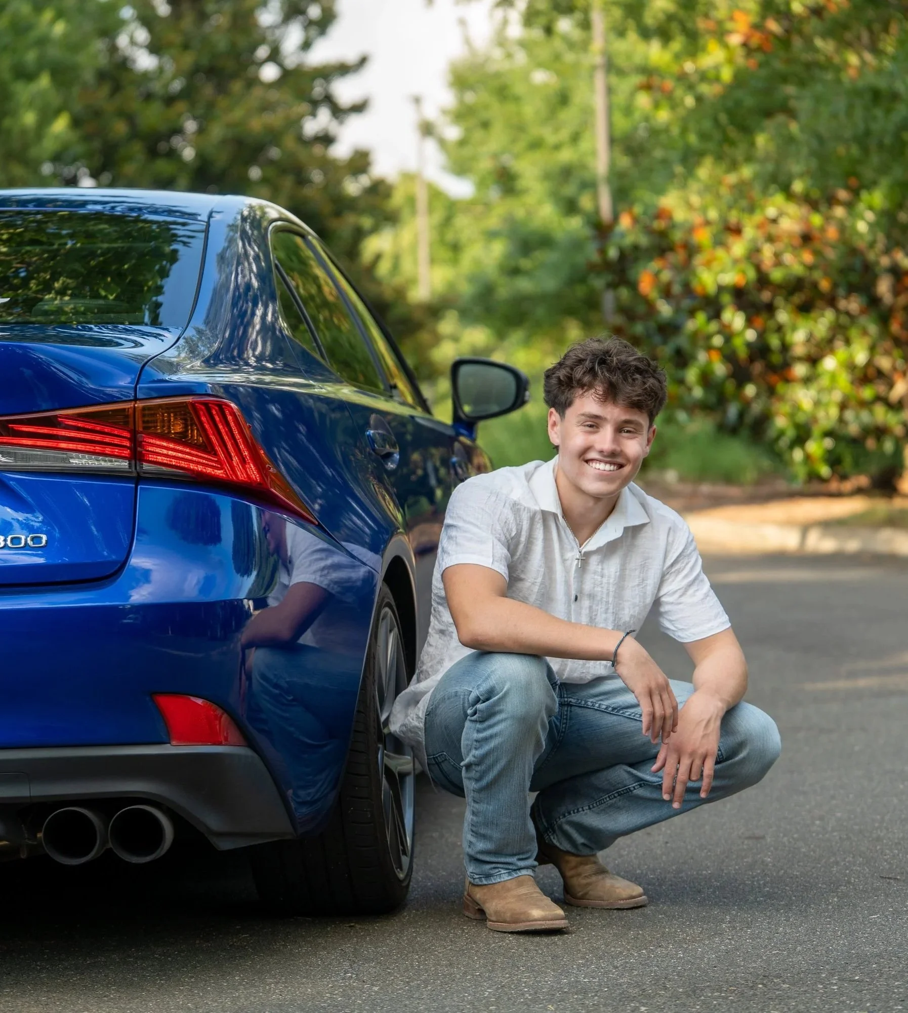 A young man in a white top and blue jeans poses against green foliage squating against his blue Lexus at E. Carroll Joyner Park, Wake Forest, North Carolina for High School Senior Photos