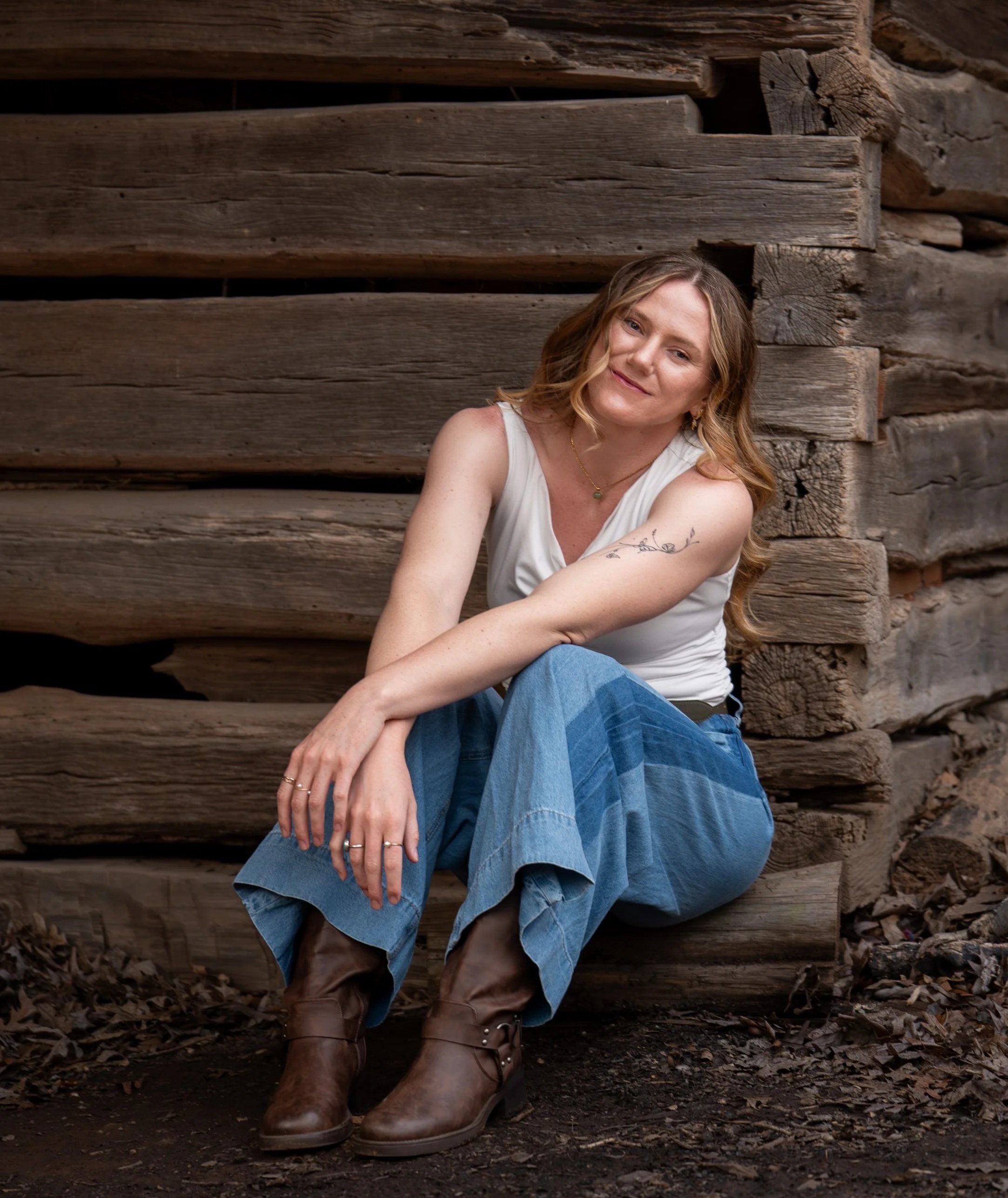 A young woman in a white top and blue jeans poses in front of a rustic wood mill at West Point on the Eno Park, Durham North Carolina for High School Senior Photos