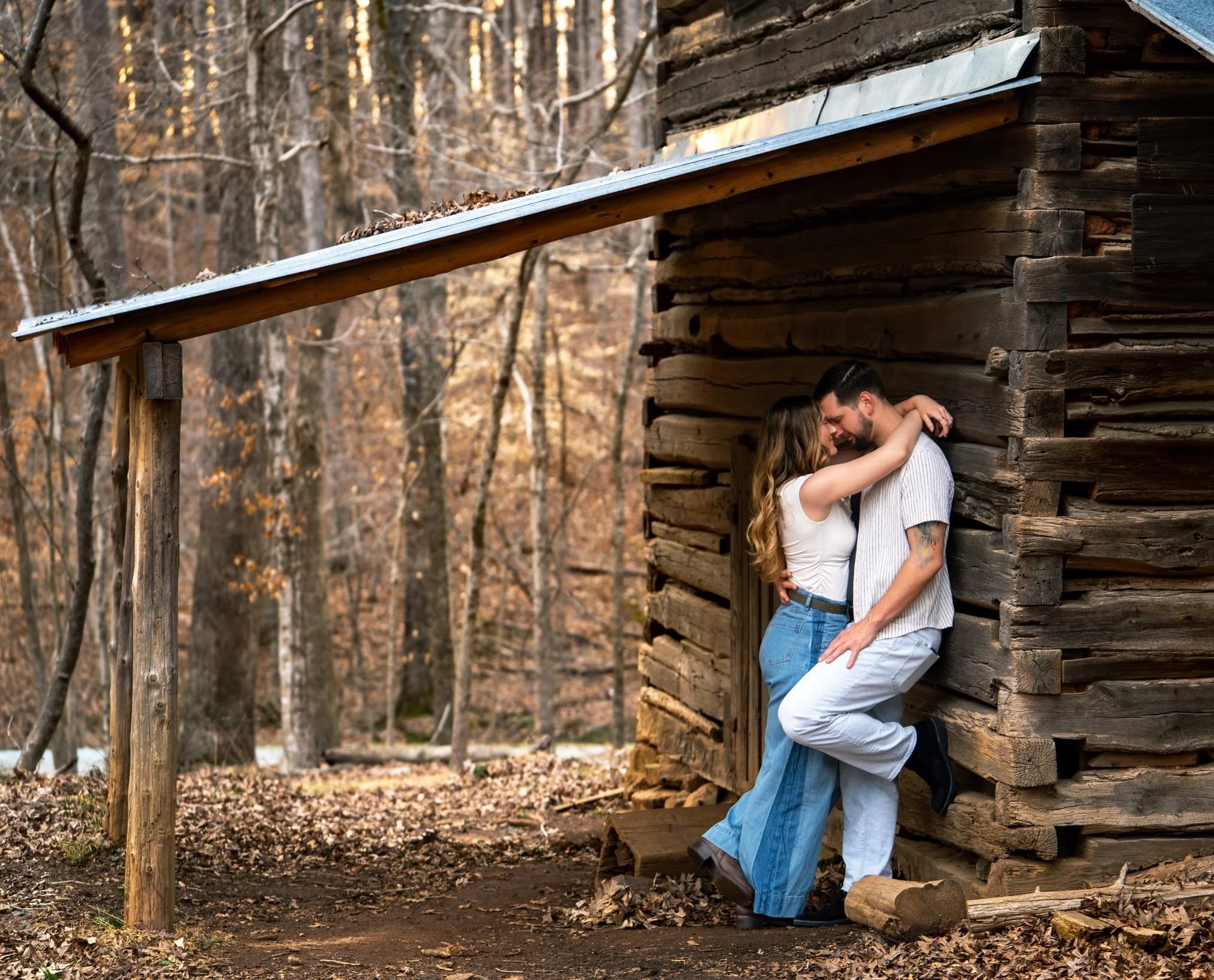 A young man & woman pose in a romantic & loving pose both in white tops and blue jeans in front of a rustic wood mill at West Point on the Eno Park, Durham North Carolina for their couple photos.
