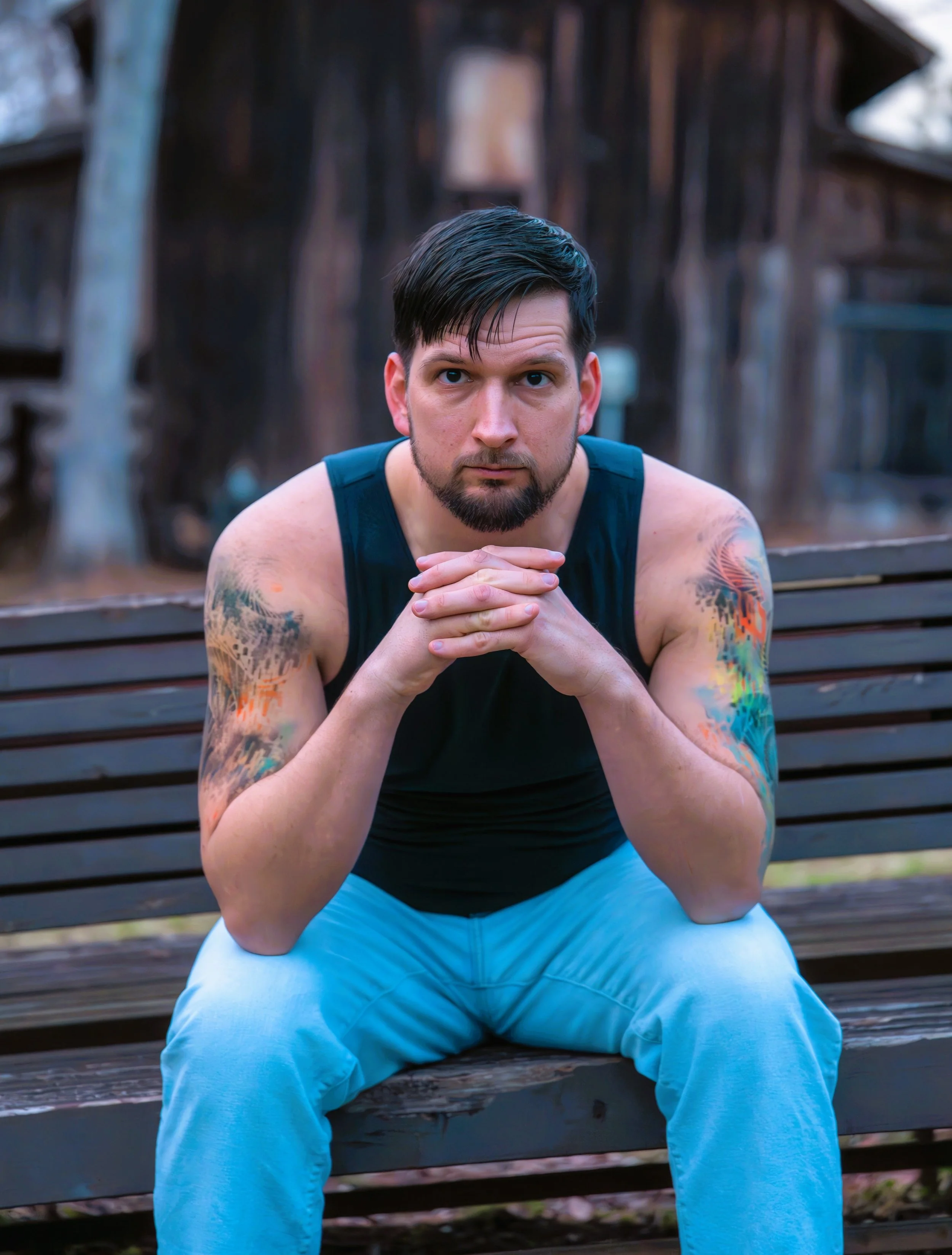 A young man in a black shirt, blue jeans. and tattoos poses in front of a rustic wood mill at West Point on the Eno Park, Durham North Carolina for Portrait Photography