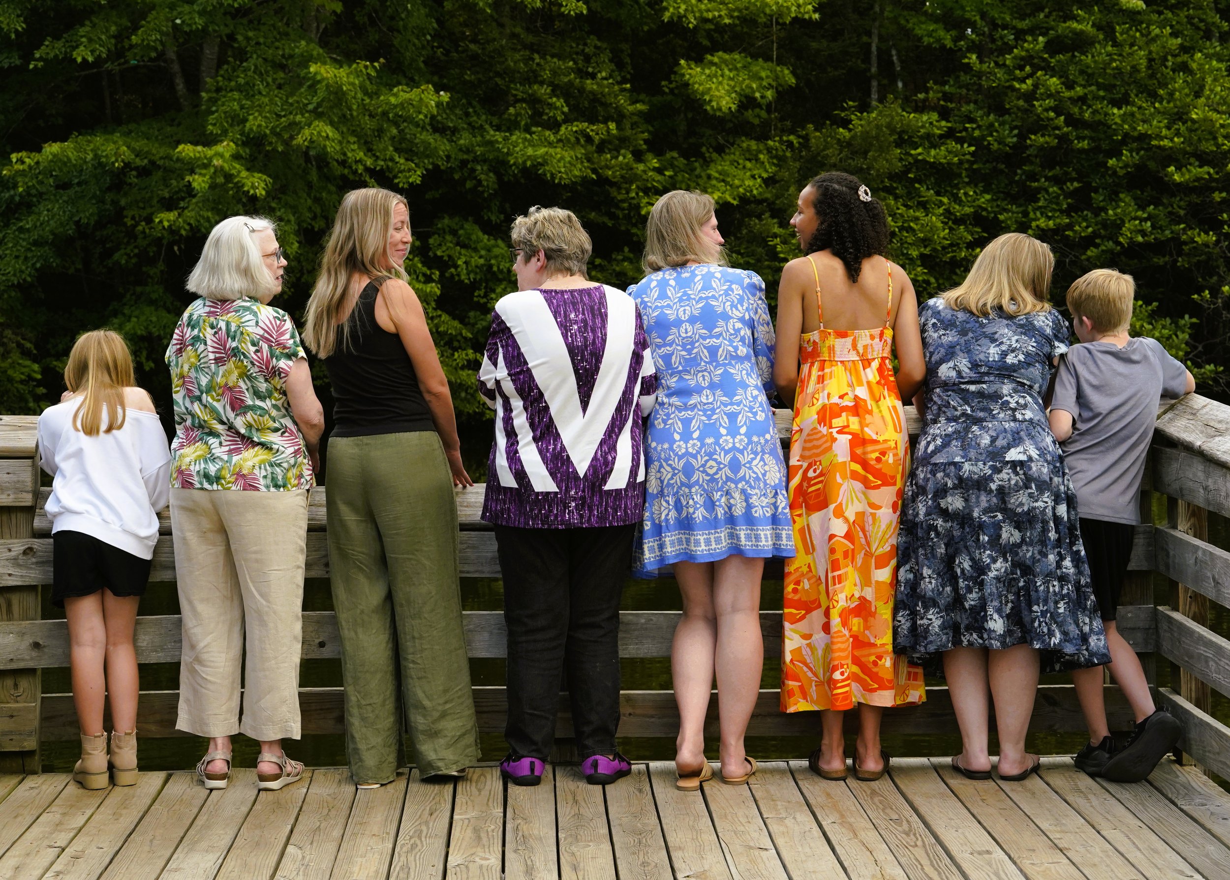 A big family photography photo of eight people - from grandma to grandchildren - wearing a mix of colors and floral patterns against a background of trees on a wooden dock at Seagroves Park, Apex, North Carolina