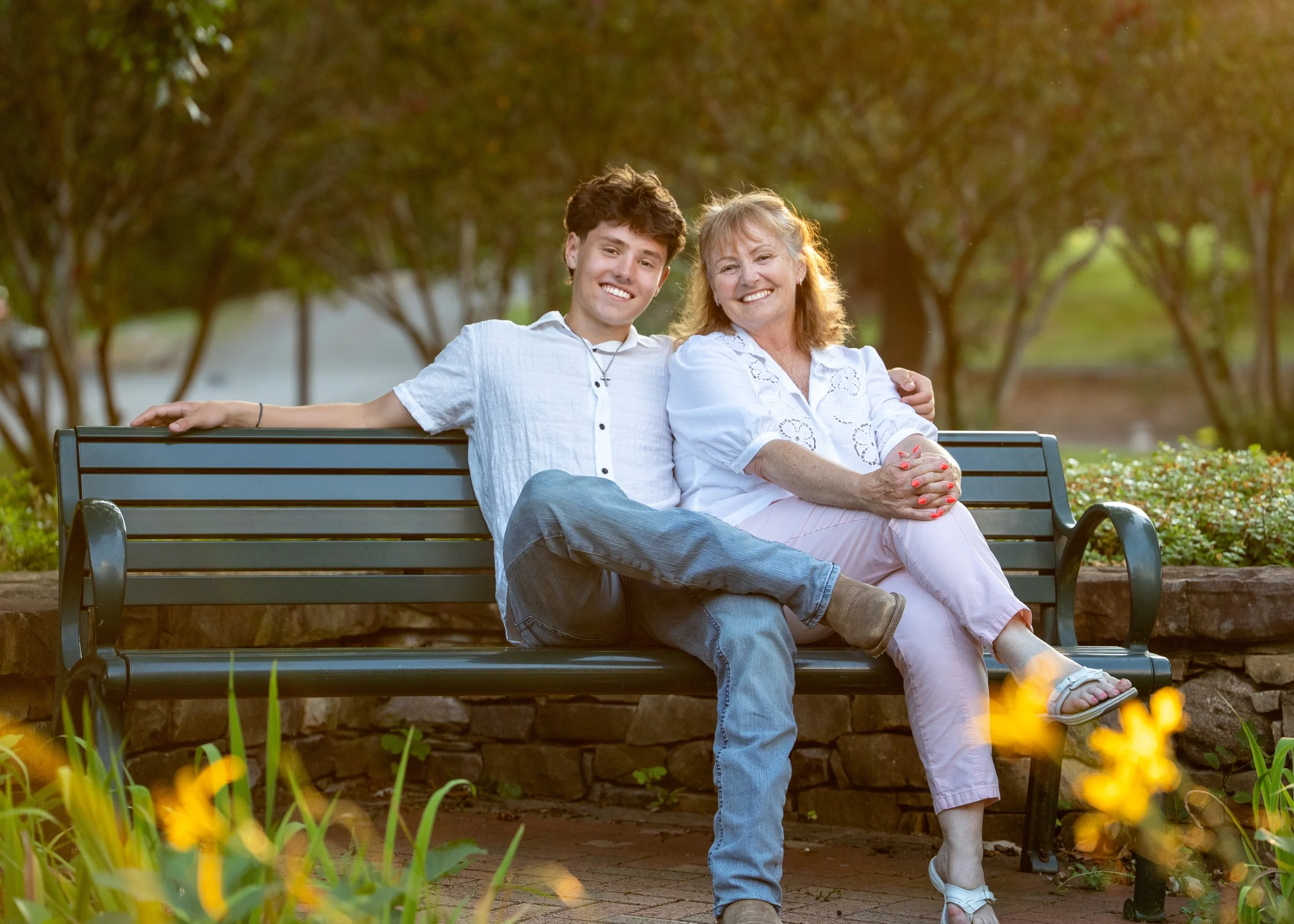 A young man in a white top and blue jeans sits with his mother on a bench with the beautiful flowers and foliage in front and the golden hour sunset behind at E. Carroll Joyner Park, Wake Forest, North Carolina for High School Senior Photos