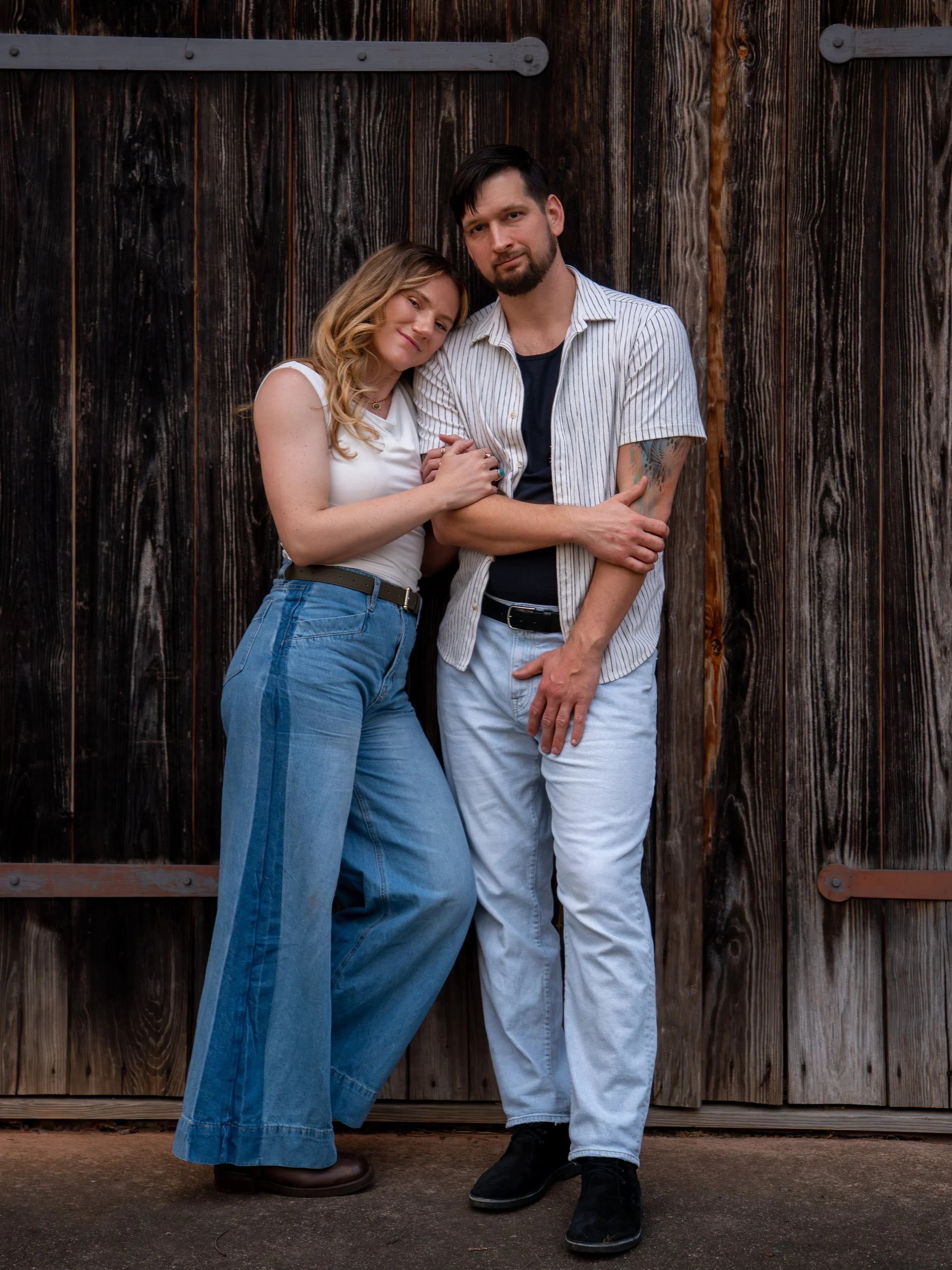 A young man & woman pose in a romantic & loving pose both in white tops and blue jeans in front of a rustic wood mill at West Point on the Eno Park, Durham North Carolina for their couple photos.