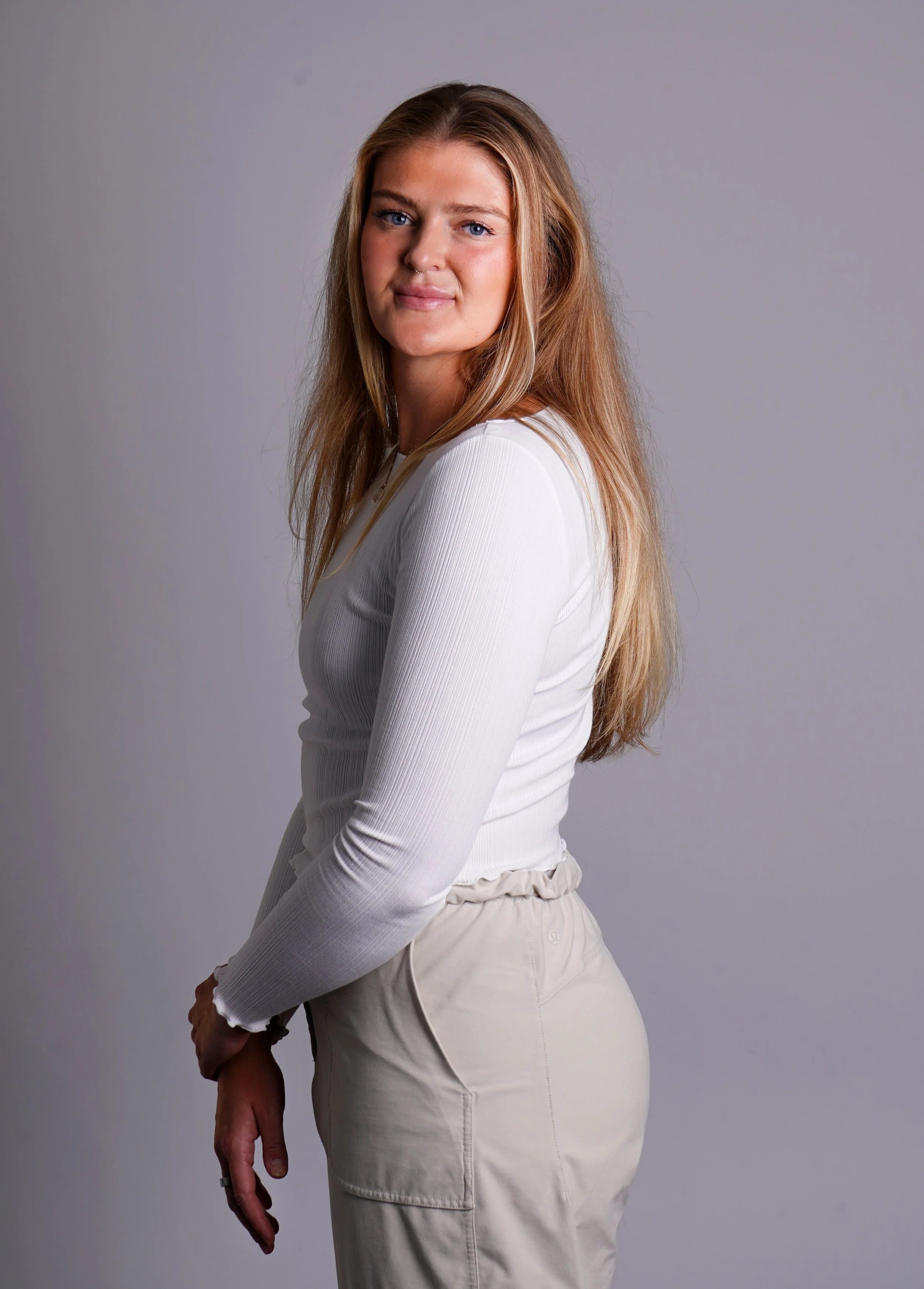A young woman with long, wavy blonde hair and blue eyes wearing a white long-sleeve top and beige pants, standing against a plain gray background in Raleigh Photography Studio