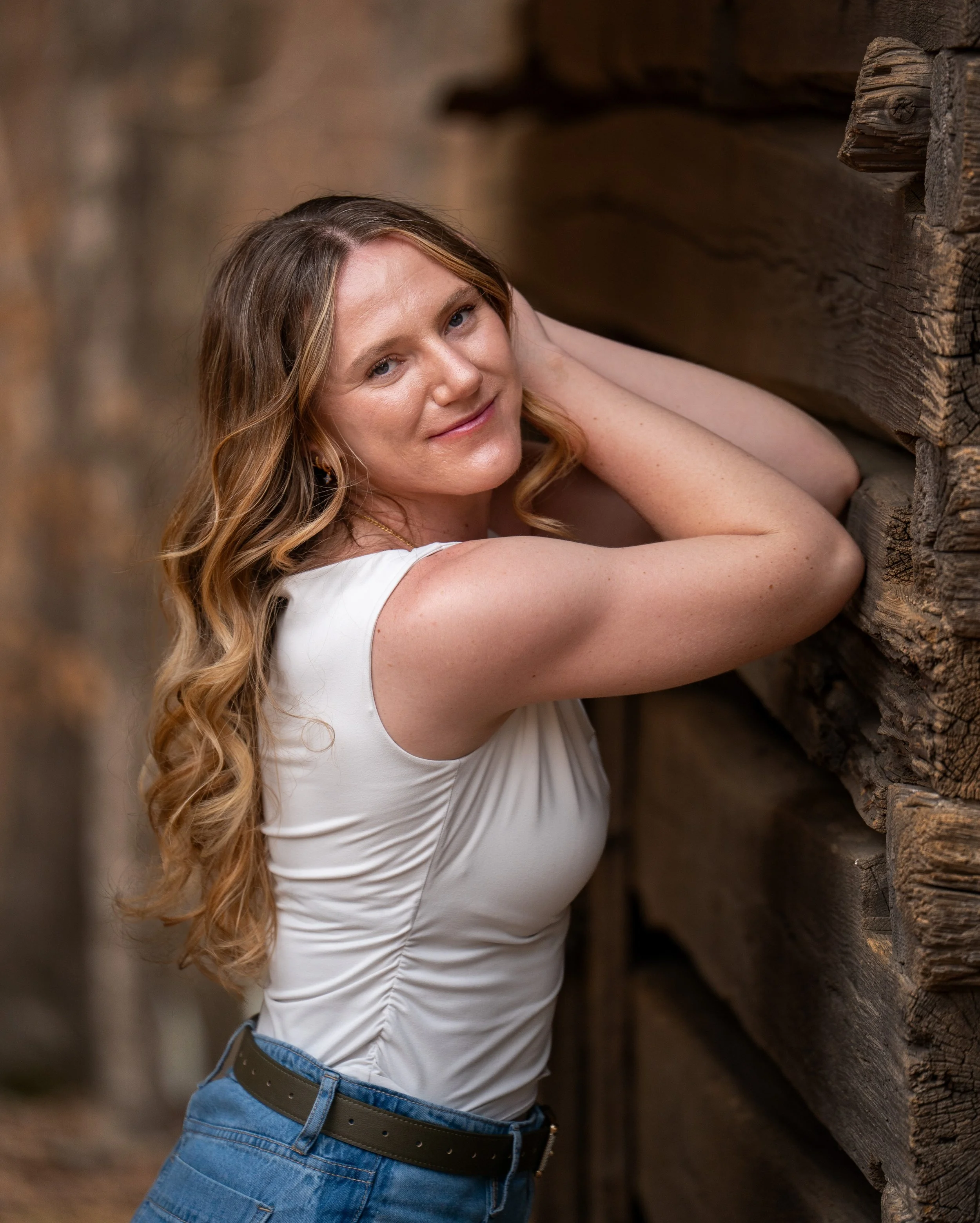 A young woman in a white top and blue jeans poses in front of a rustic wood mill at West Point on the Eno Park, Durham North Carolina for High School Senior Photos