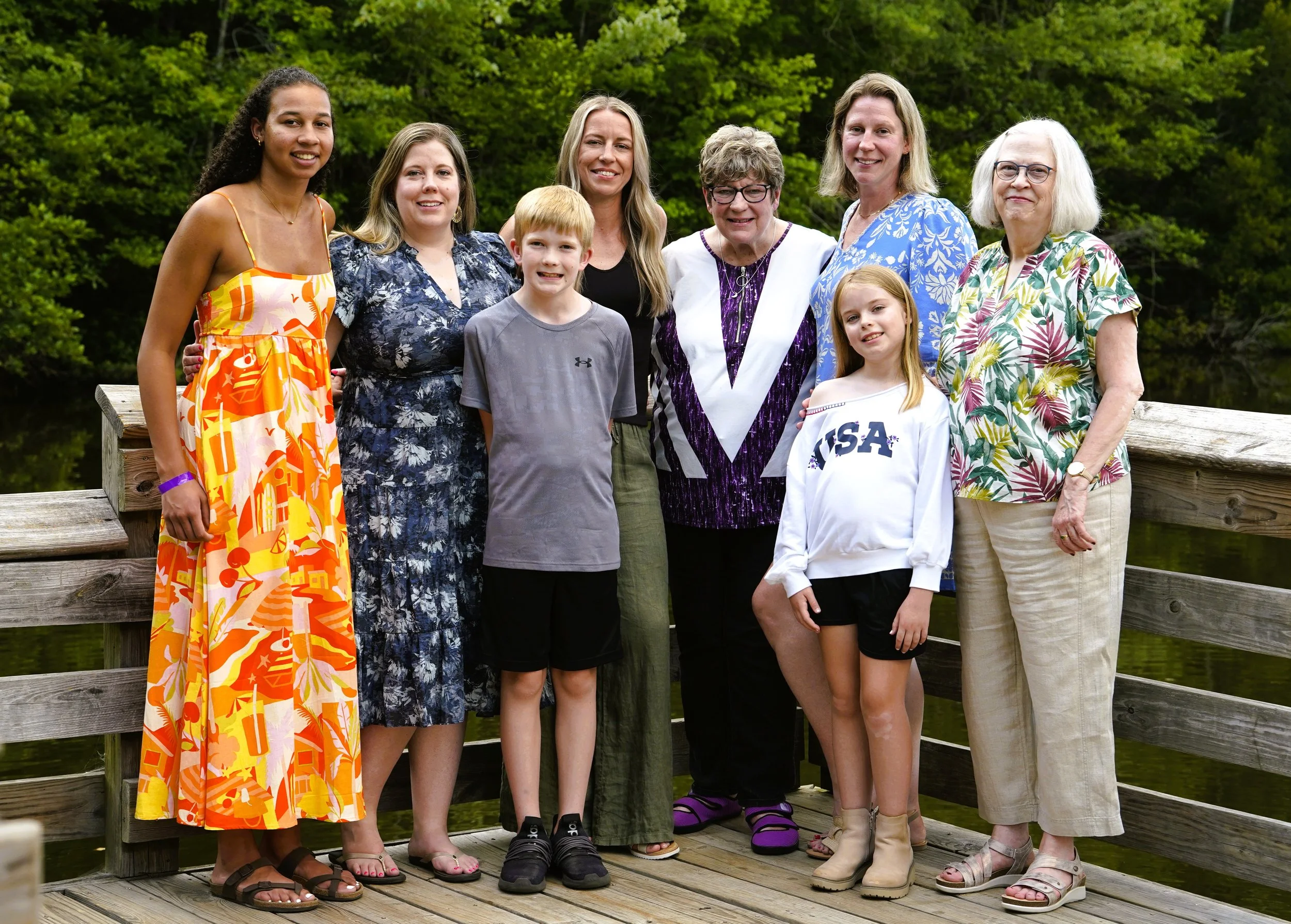 A big family photography photo of eight people - from grandma to grandchildren - wearing a mix of colors and floral patterns against a background of trees on a wooden dock at Seagroves Park, Apex, North Carolina