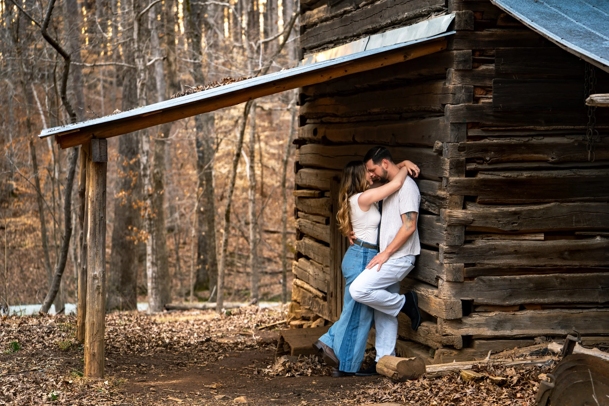 A young man & woman pose in a romantic & loving pose both in white tops and blue jeans in front of a rustic wood mill at West Point on the Eno Park, Durham North Carolina for their couple photos.