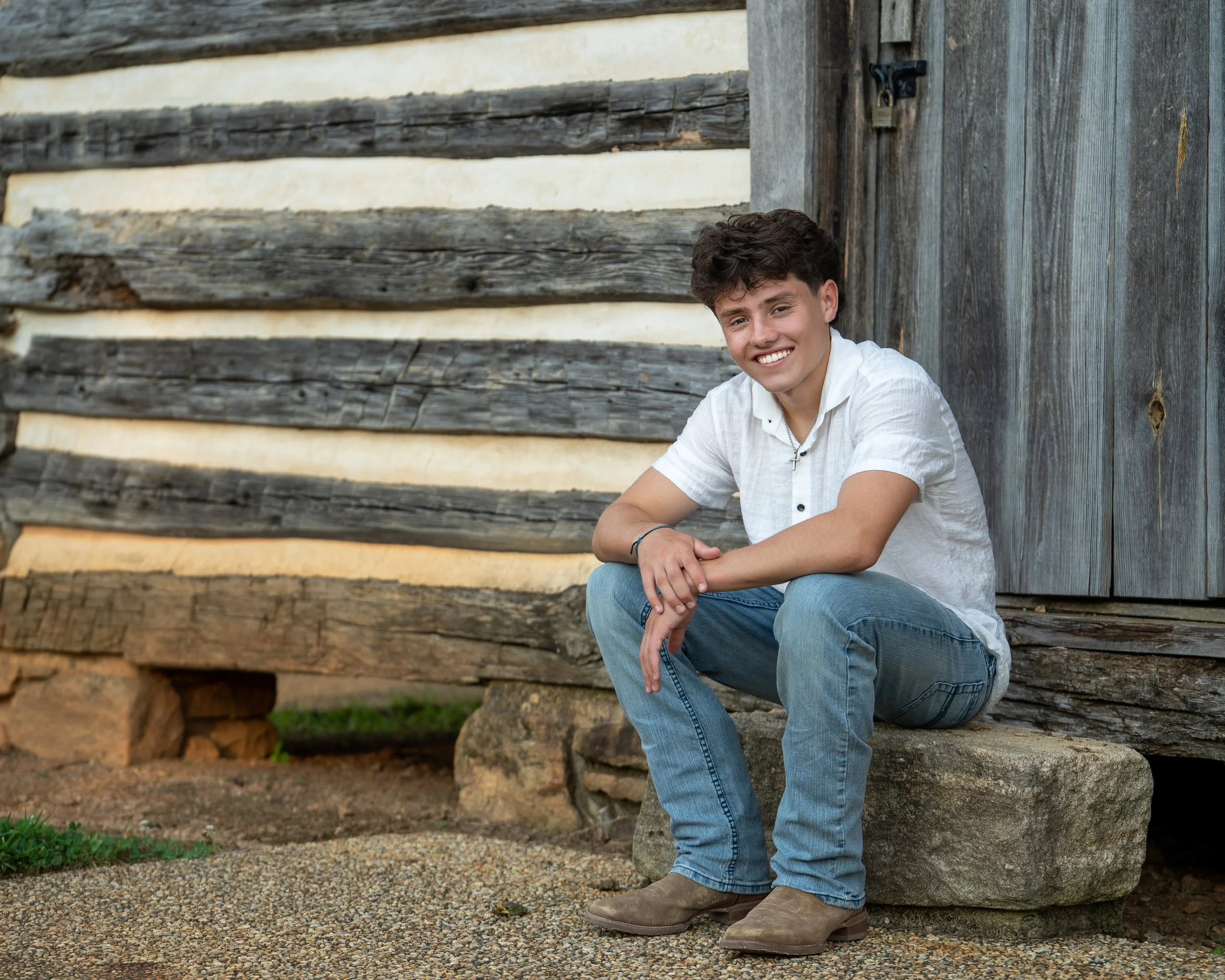 A young man in a white top and blue jeans squats on a large rock in front of a rustic shed at E. Carroll Joyner Park, Wake Forest, North Carolina for High School Senior Photos