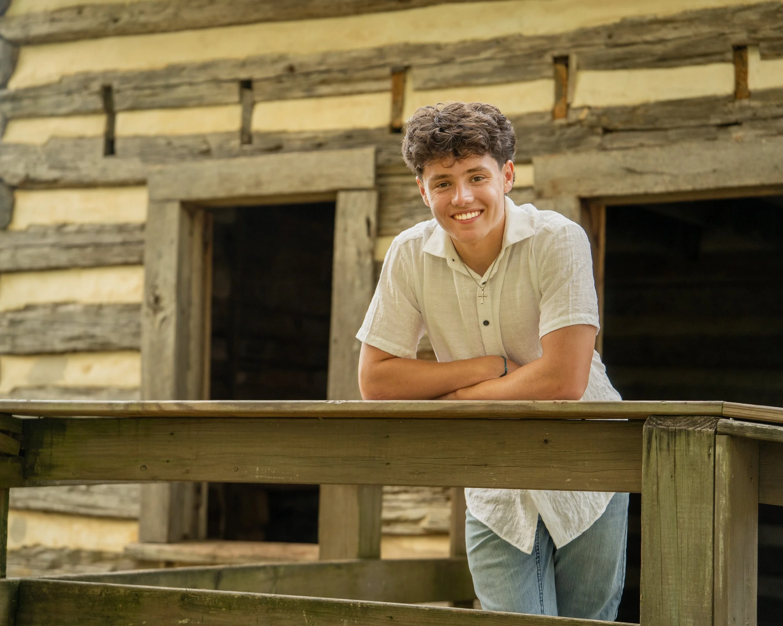 A young man in a white top and blue jeans poses against a wood railing in front of a rustic shed at E. Carroll Joyner Park, Wake Forest, North Carolina for High School Senior Photos