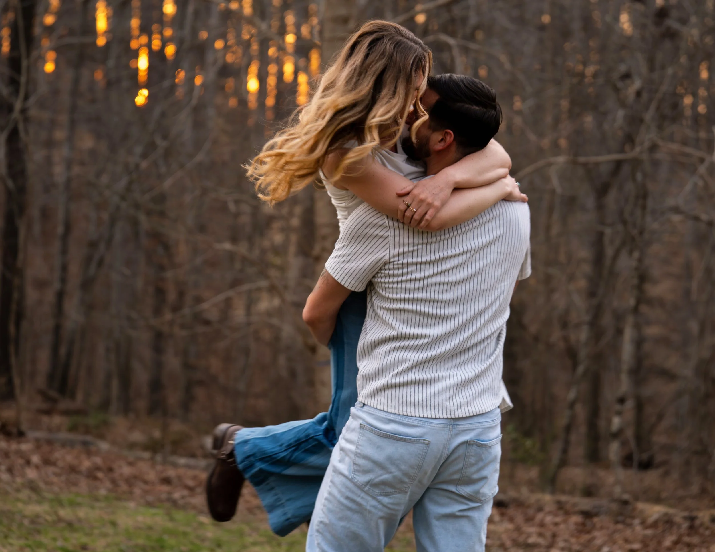 A young man picks up and spins his girlfriend in a romantic & fun pose both in white tops and blue jeans in front of a fire orange sunset at West Point on the Eno Park, Durham North Carolina for their couple photos.