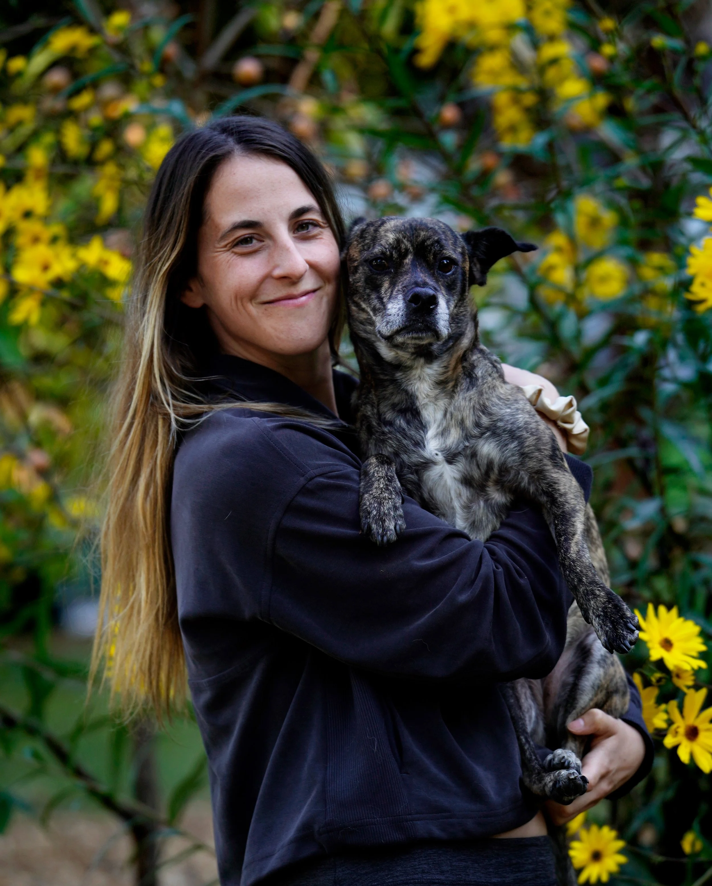 A woman with long brown hair smiling while holding a small brindle dog outdoors with yellow swamp sunflowers in the background.